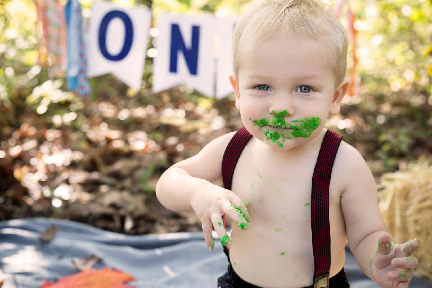 Toddler boy with green cake smash frosting on face wearing suspenders at outdoor birthday.
