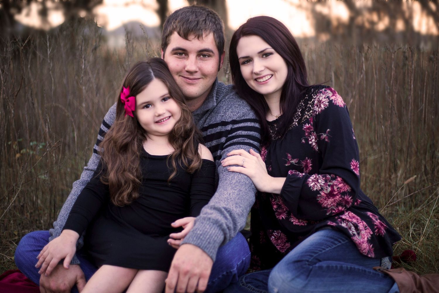 A smiling family of three poses outdoors in a field, with a young girl in a black dress and her parents.