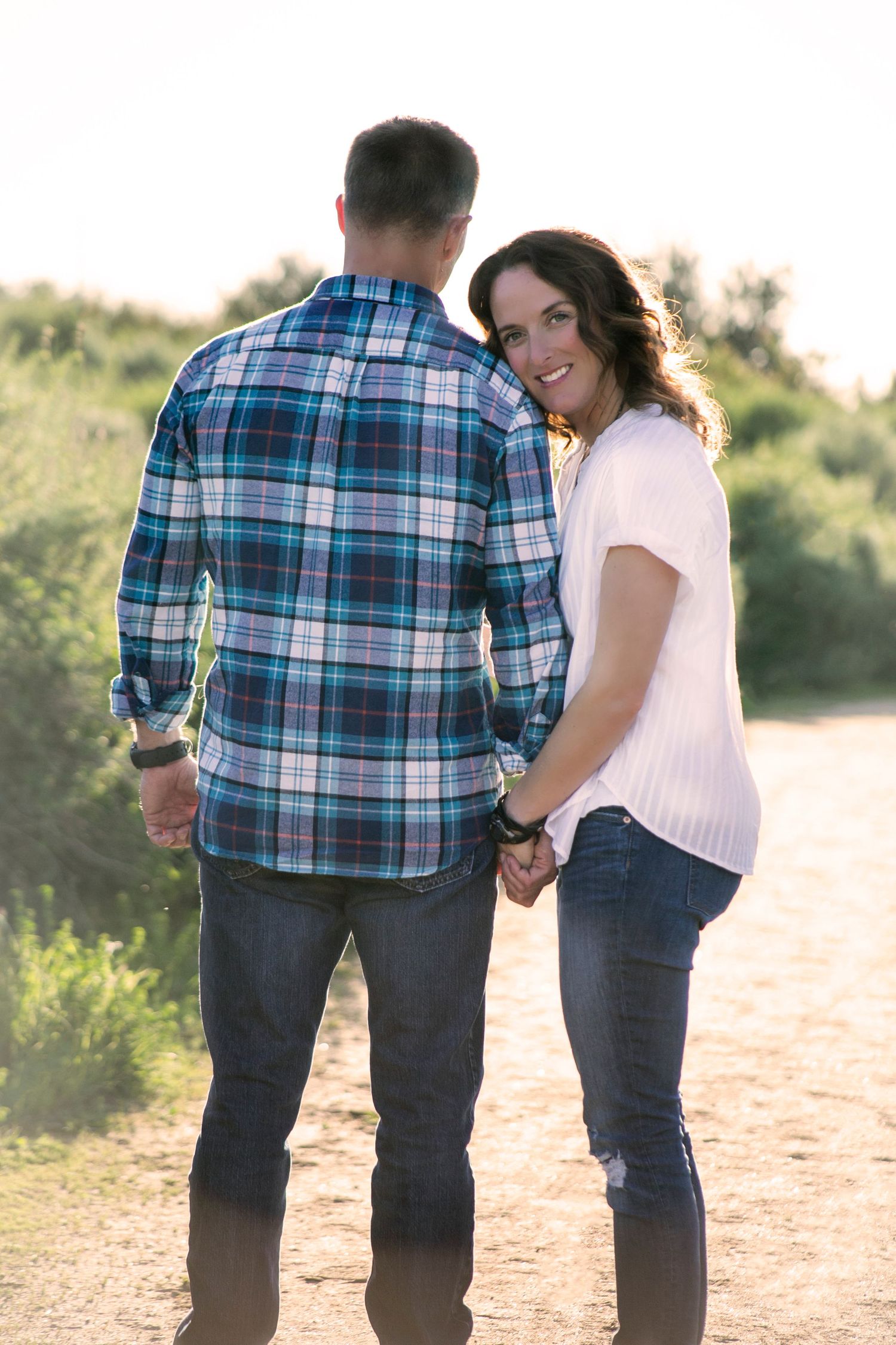 Couple holding hands on dirt path at sunset, woman smiling back at camera during couples session.