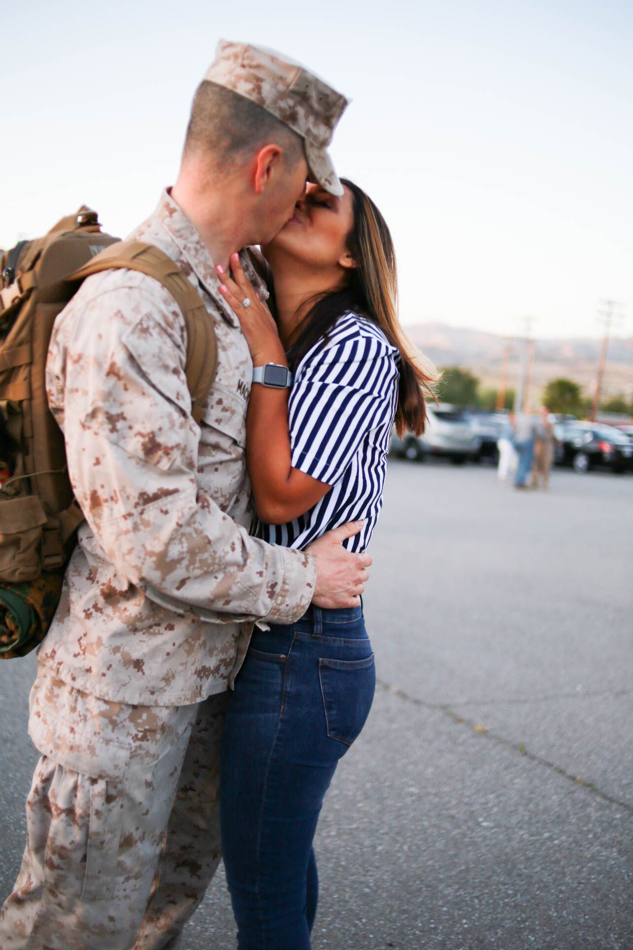Military soldier in camouflage uniform kissing partner in striped shirt and jeans in outdoor parking lot.