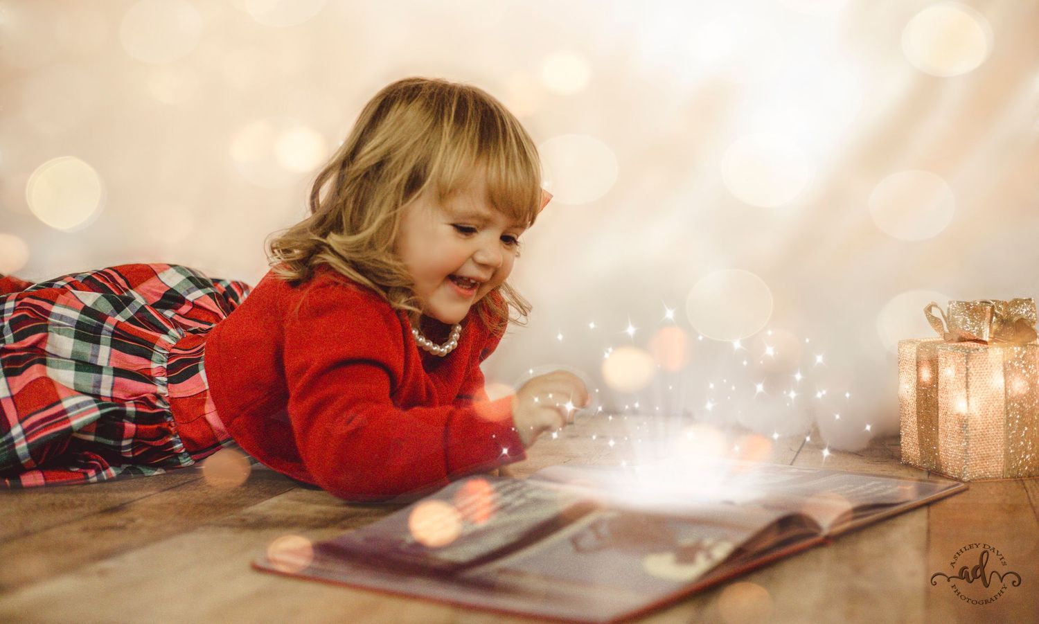 Toddler girl in red dress reading a magical glowing book with bokeh Christmas lights.