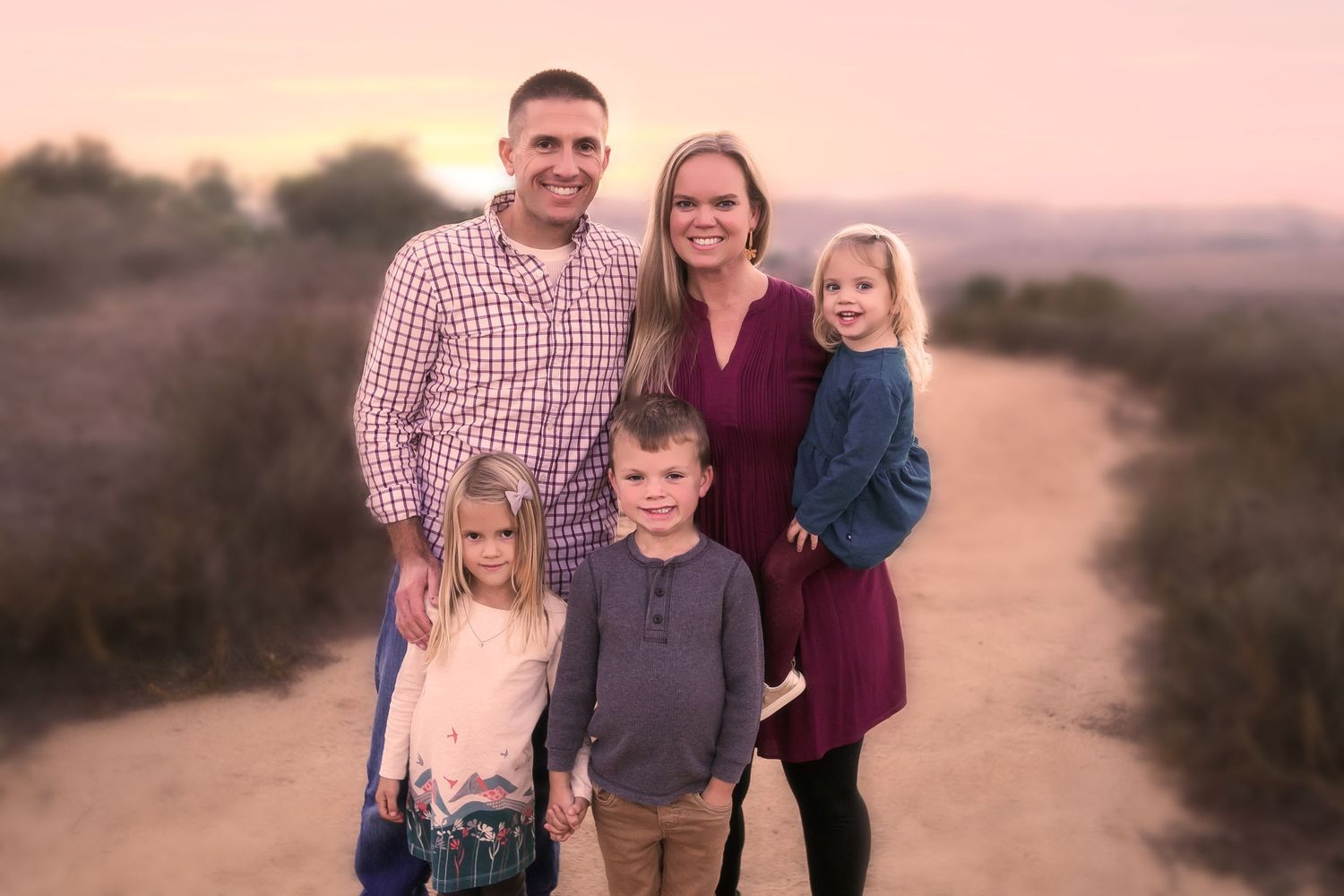 Happy family of five poses together on dirt path during golden hour sunset.