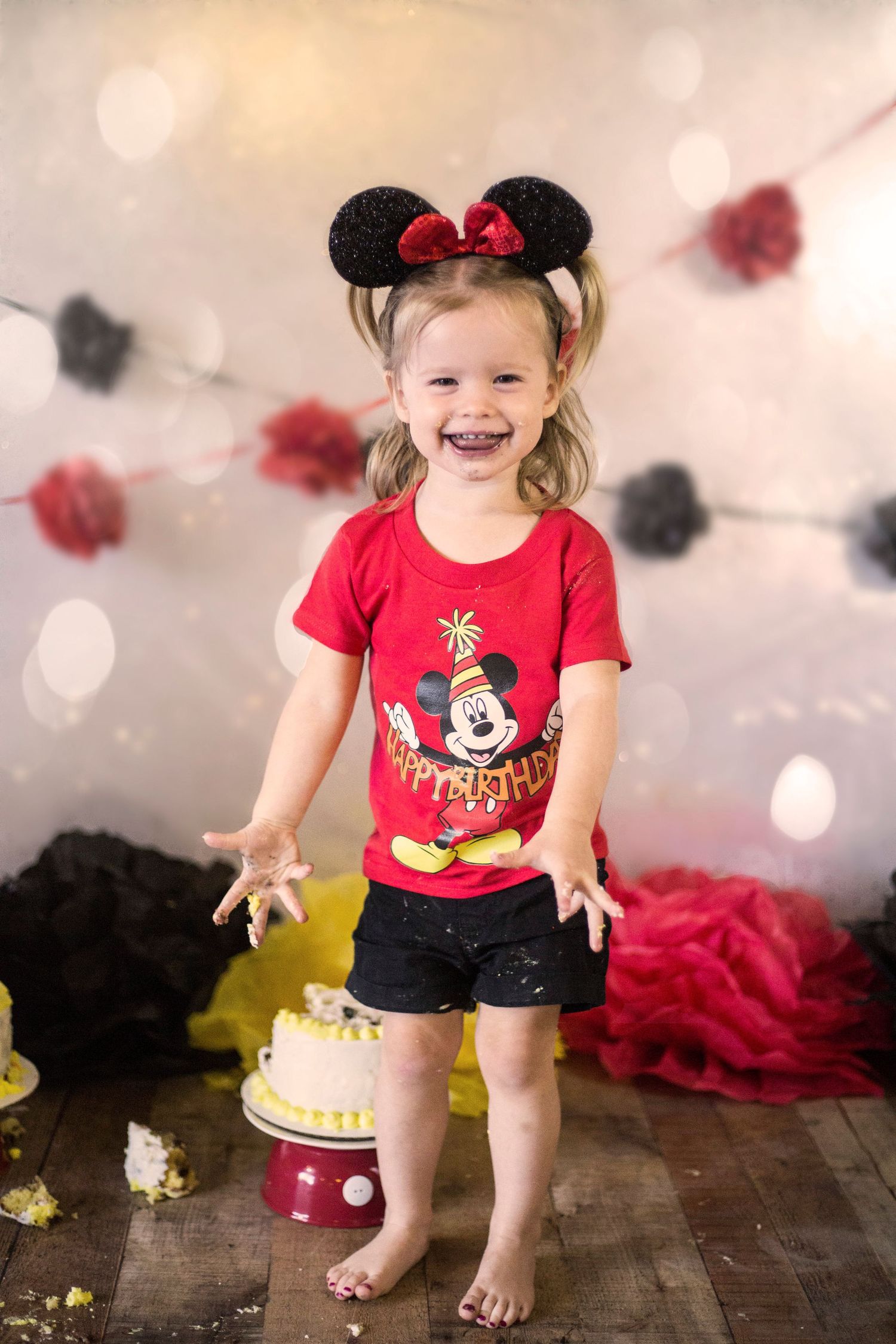 Happy toddler girl in Minnie Mouse ears and red shirt at a cake smash birthday party.