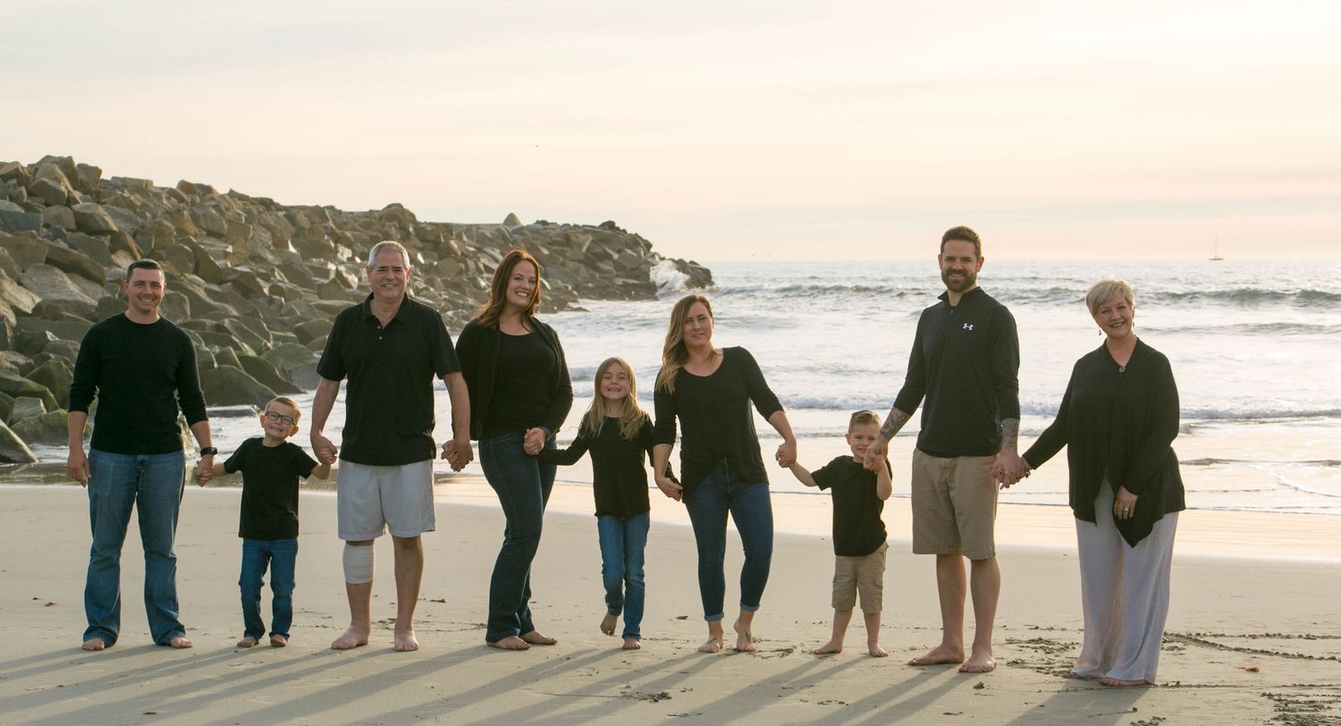 Extended family of eight poses together on rocky beach at sunset in matching black outfits.