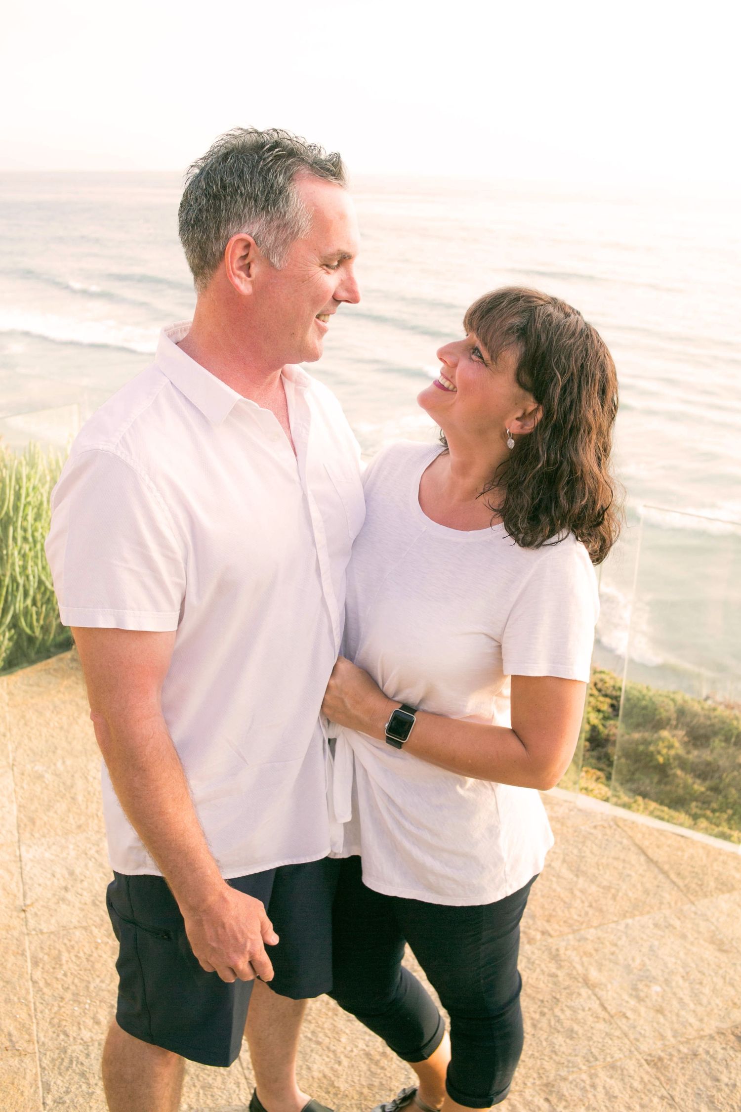 Happy mature couple in white shirts smiling at each other with ocean view in background.