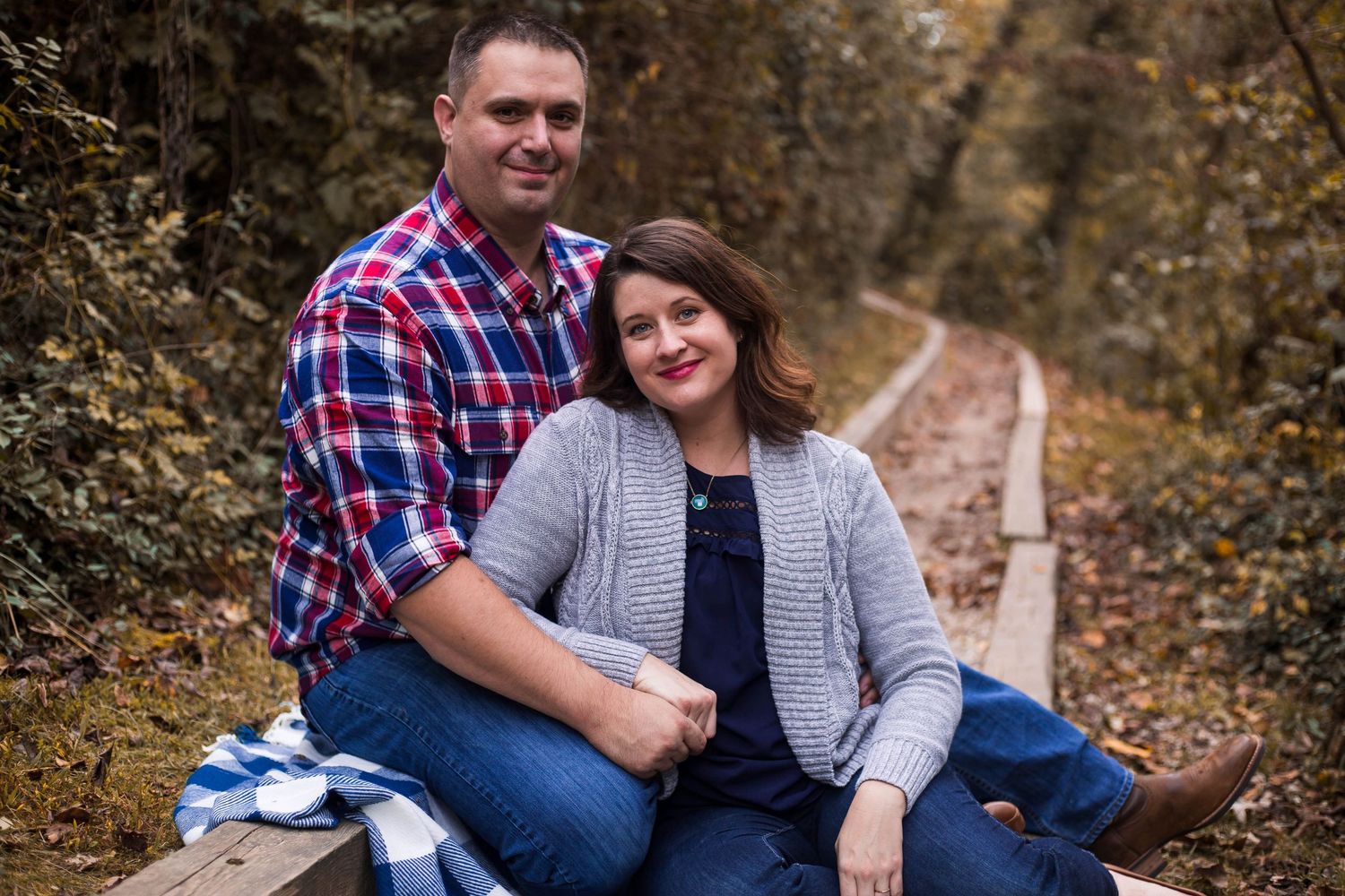 Couple sitting together on wooden path surrounded by autumn foliage wearing casual plaid and gray.