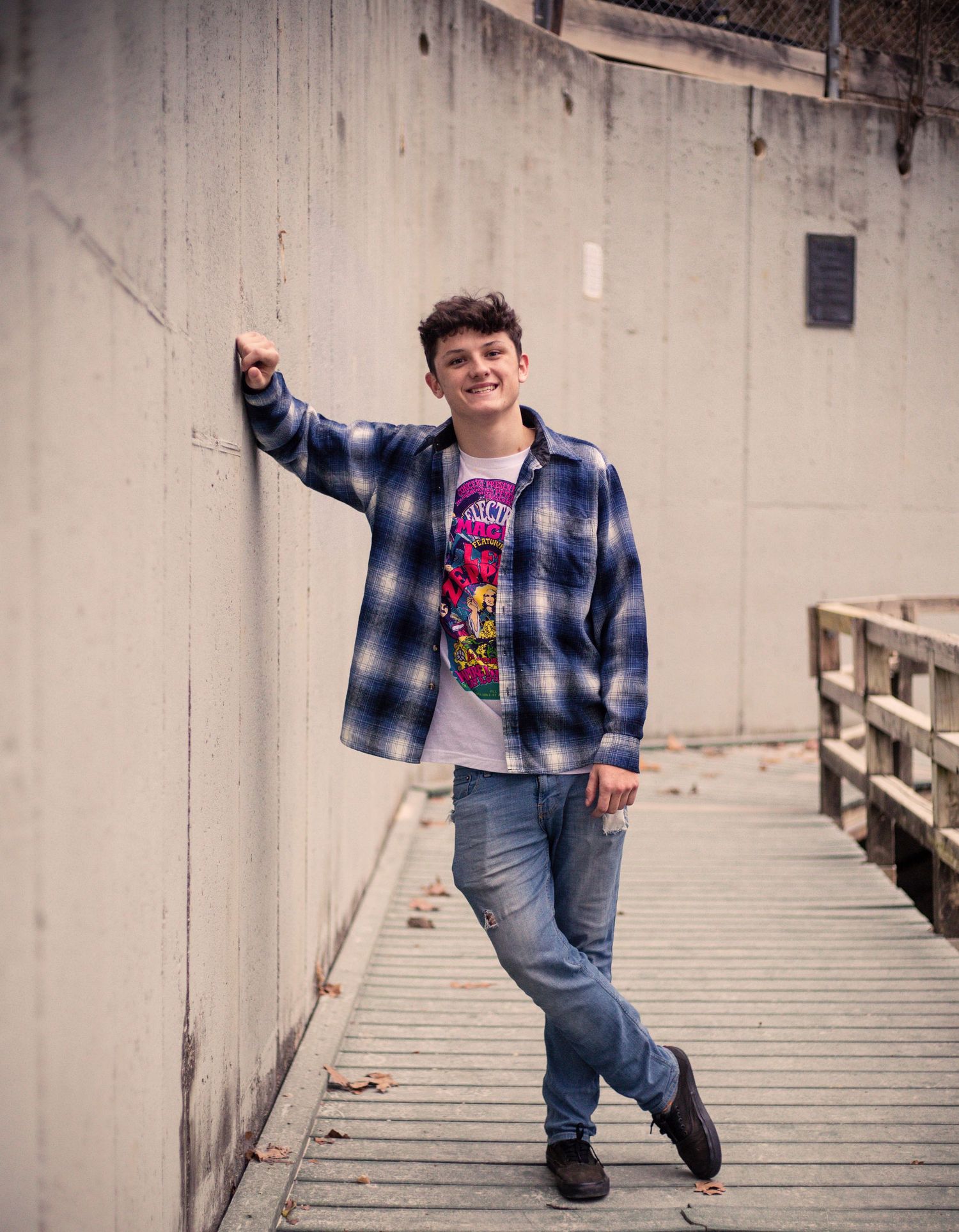 Teen boy in plaid flannel shirt leaning against wall on wooden bridge for senior portraits.