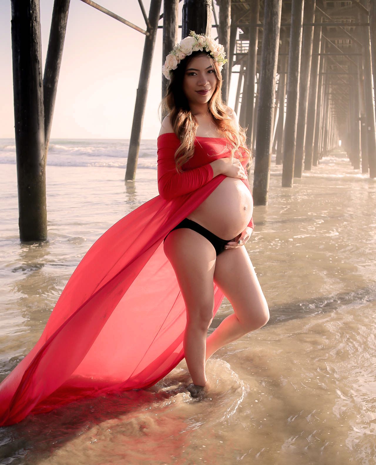 Pregnant woman in red flowing fabric poses under pier at beach during sunset.