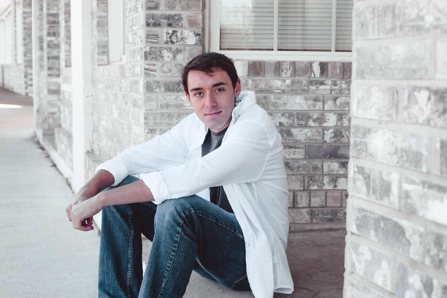 Young man in white coat sitting against brick wall outdoors for senior portrait photography session.