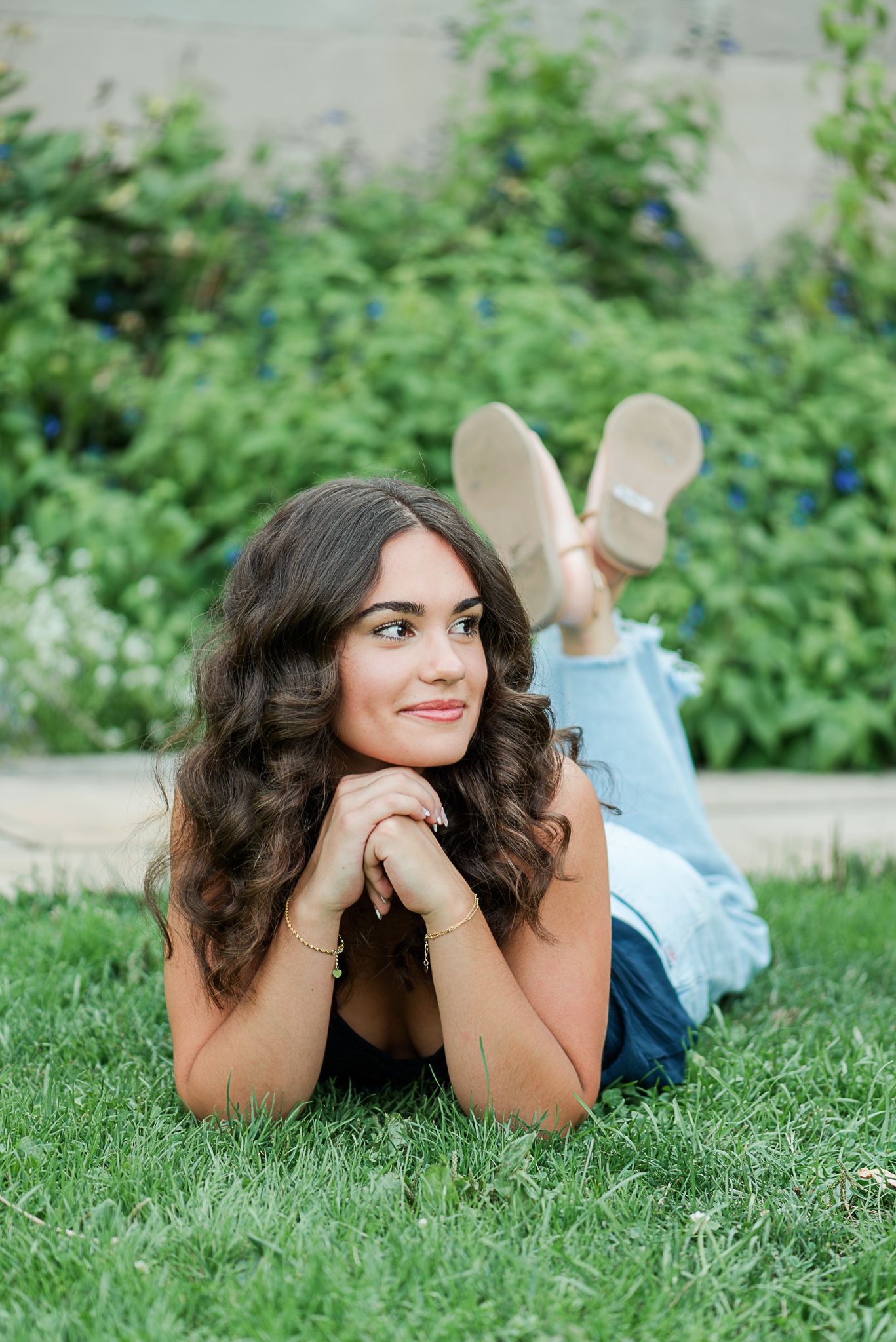 A young woman in casual attire relaxes on green grass while laying on her stomach, with lush greenery in the background.