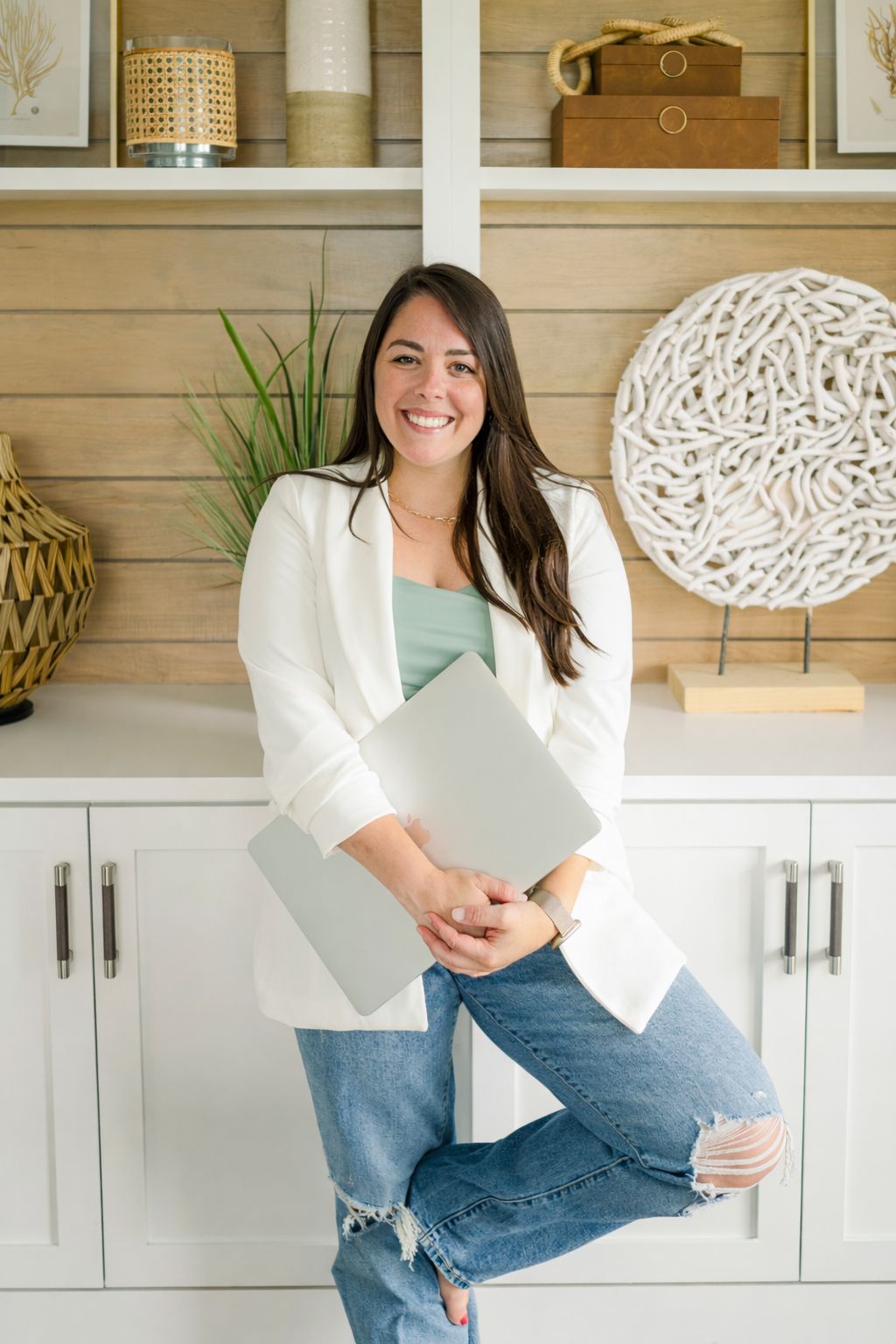A casual lifestyle photoshoot showing someone sitting on a white cabinet wearing jeans and a white blazer in a bright room.