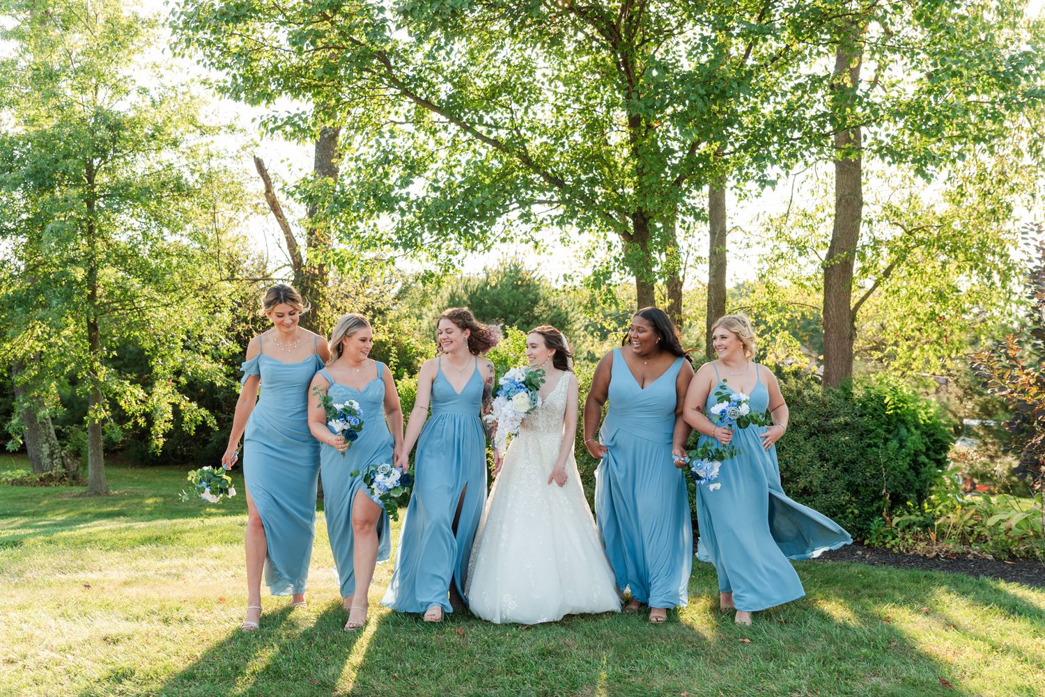 Wedding party in light blue bridesmaid dresses and a white bridal gown pose together on sunny garden lawn.
