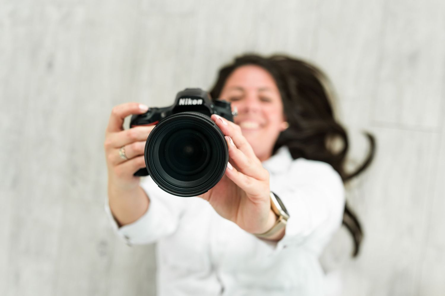 A photographer in white holds up a Nikon camera while taking photos against a light colored background.