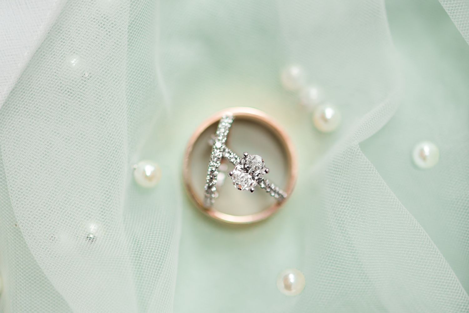 Sparkling diamond cross shaped engagement ring nestled on mint green tulle fabric with scattered white pearls.