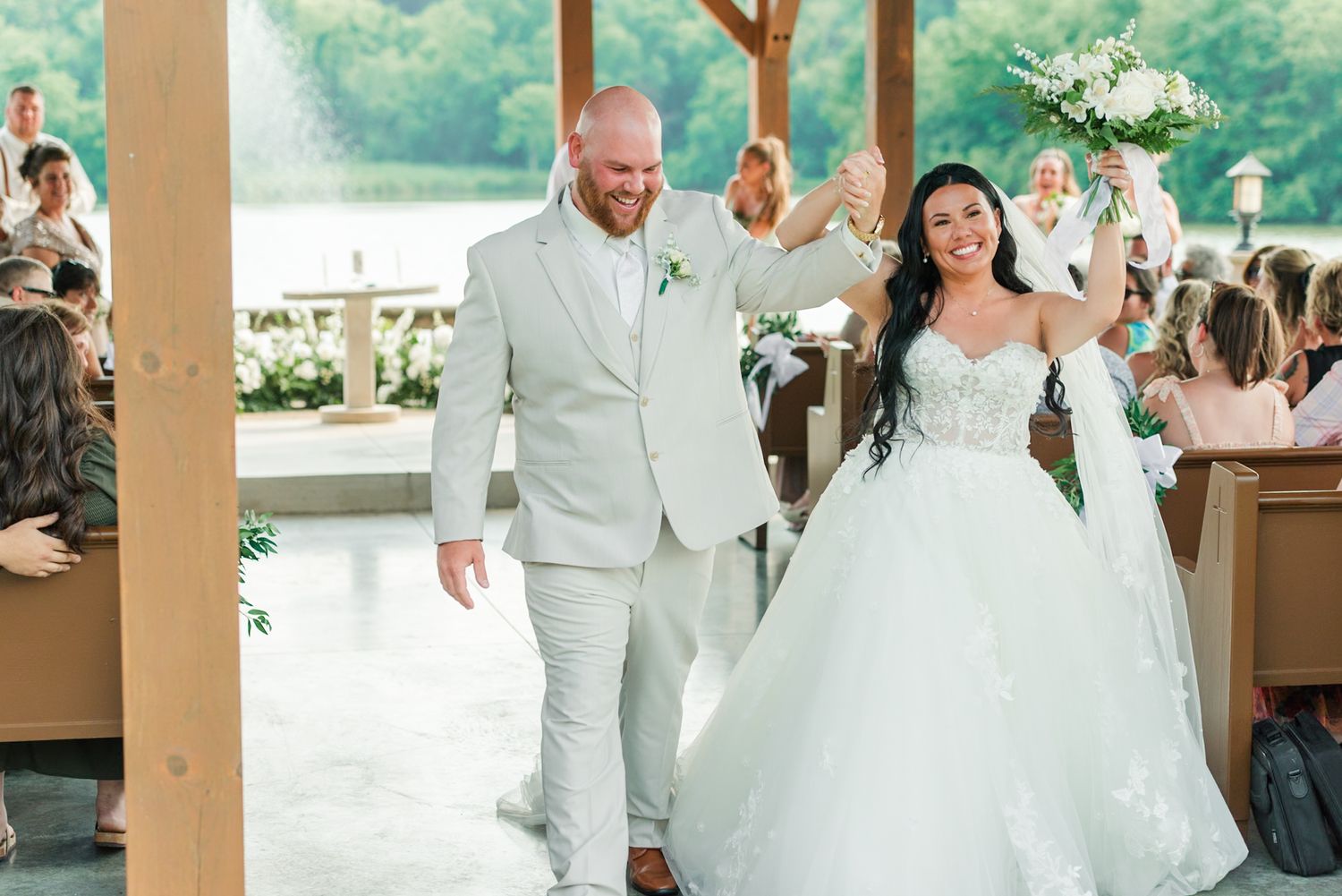 A joyful wedding celebration under a covered pavilion with the happy couple wearing white and beige attire.