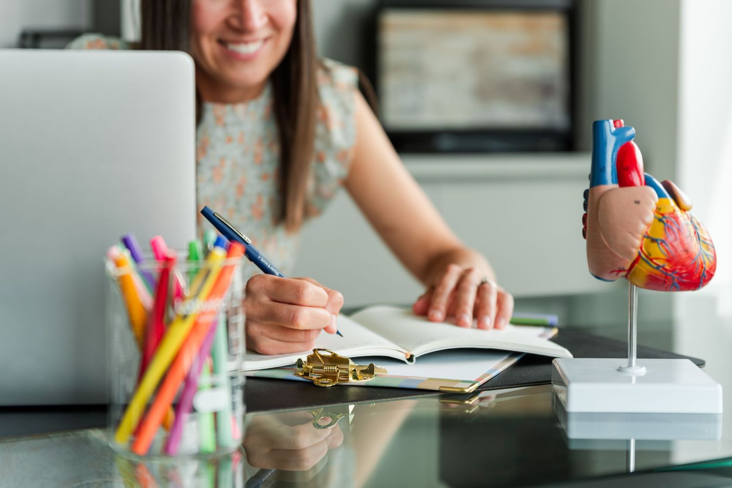 A person studying with colorful pencils and notebooks on a white desk while holding a beverage.