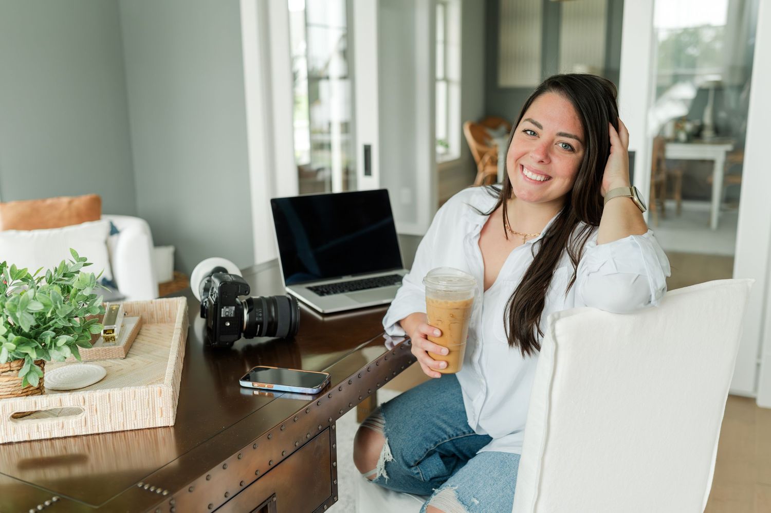 Woman in white shirt working on laptop at home office desk with coffee and plants nearby.