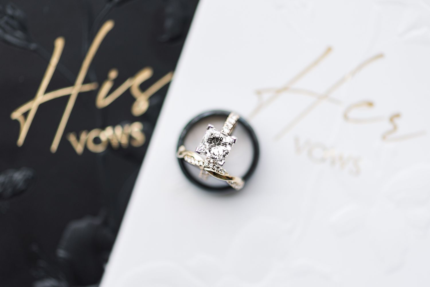 A silver heart-shaped engagement ring rests on black and gold wedding vows or cards with elegant script lettering.