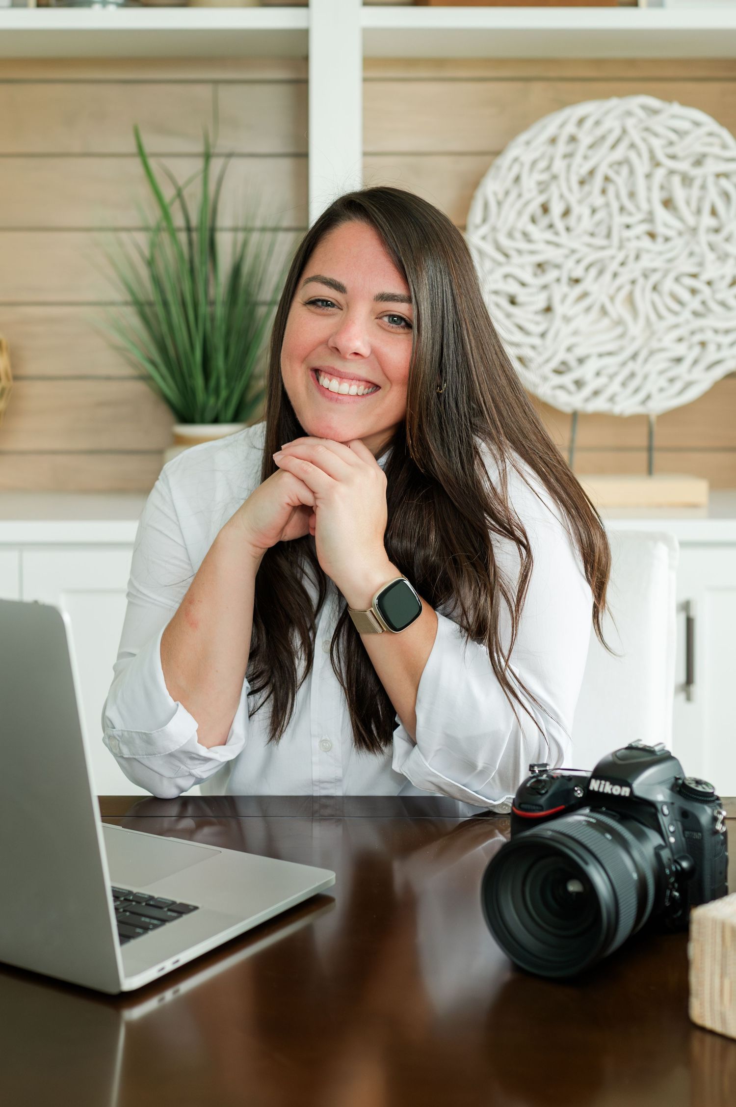 Professional photographer at desk with laptop and DSLR camera smiling in home office with modern decor.