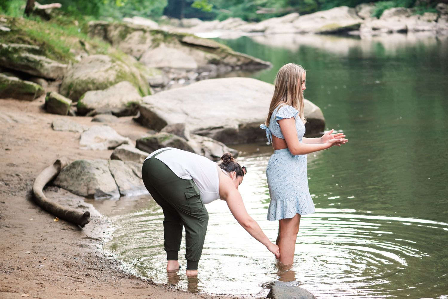 People wading and playing in a calm lake surrounded by rocks and greenery on a summer day.