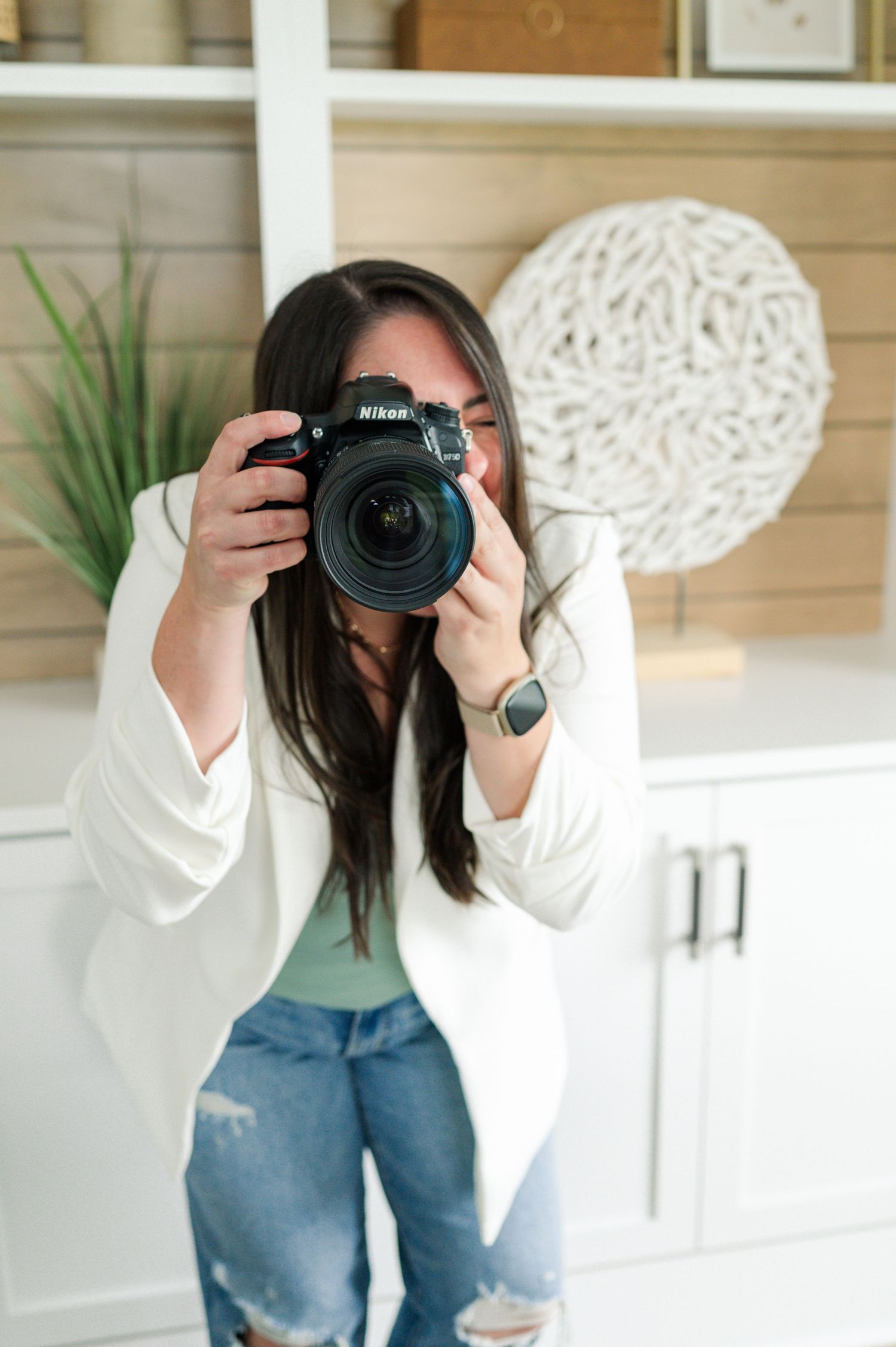 Photographer in white blazer and ripped jeans takes photo with professional camera against light wooden wall decor.