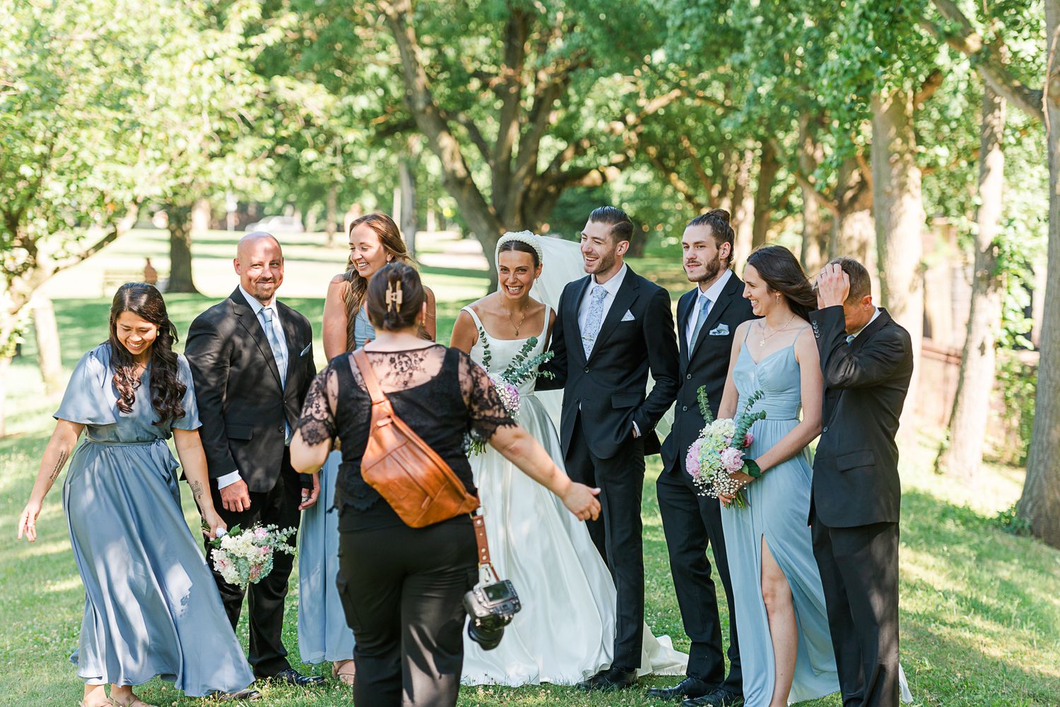 A wedding party poses together outdoors on a sunny day in a tree-lined garden setting.