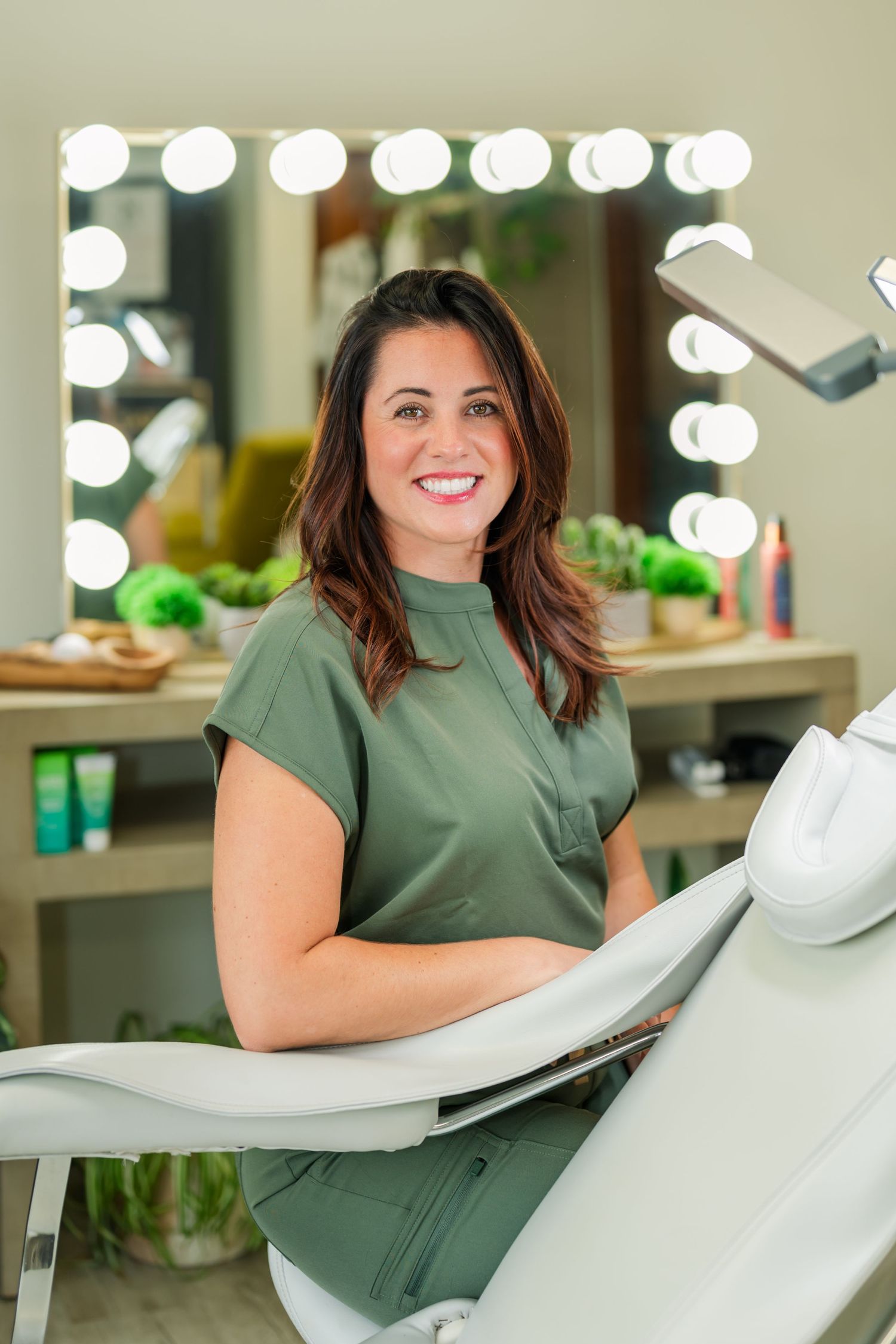 A dental professional in a sage green uniform smiles while seated in a modern treatment room with illuminated vanity lights.