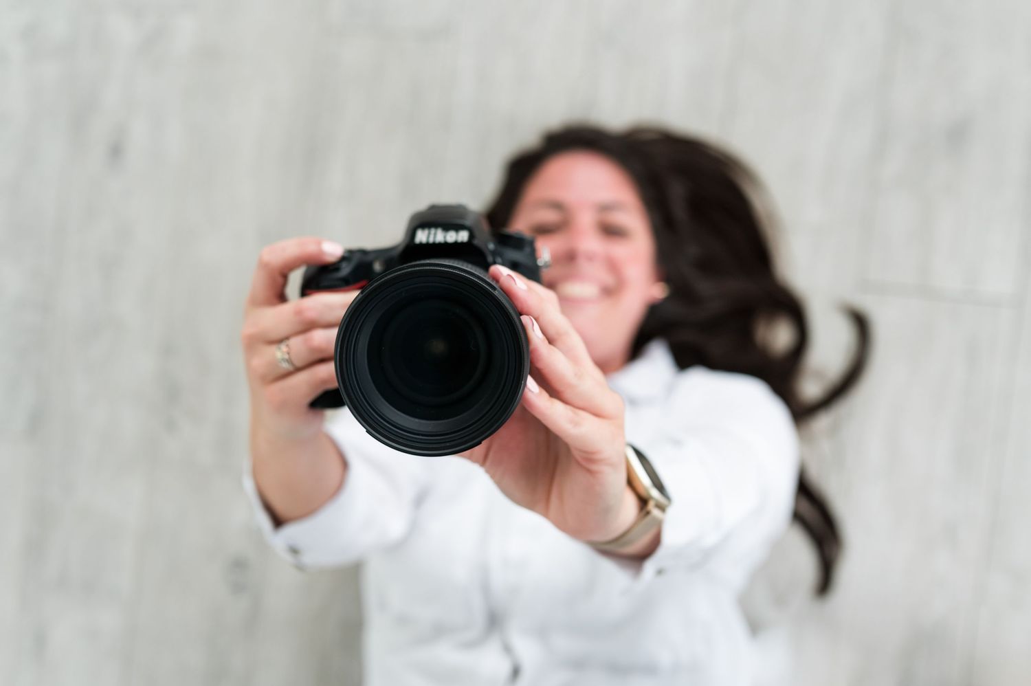 A photographer in white clothing holds up a Nikon camera lens while posing against a light colored wall.