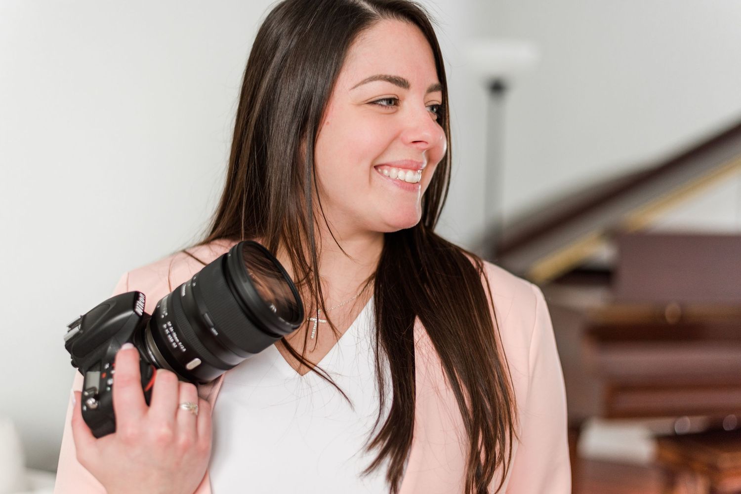 Professional photographer with long dark hair smiles while holding a DSLR camera in a bright indoor setting.
