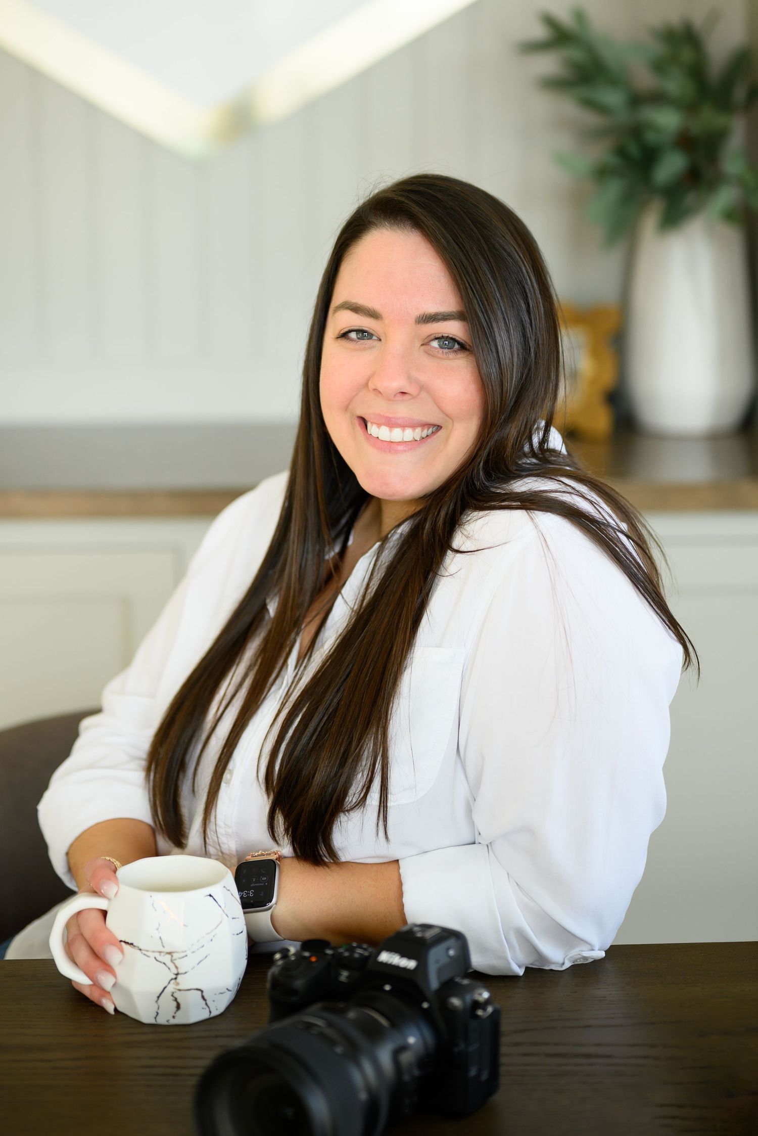 Professional photographer in white blouse smiles while sitting at wooden table with camera and coffee mug.