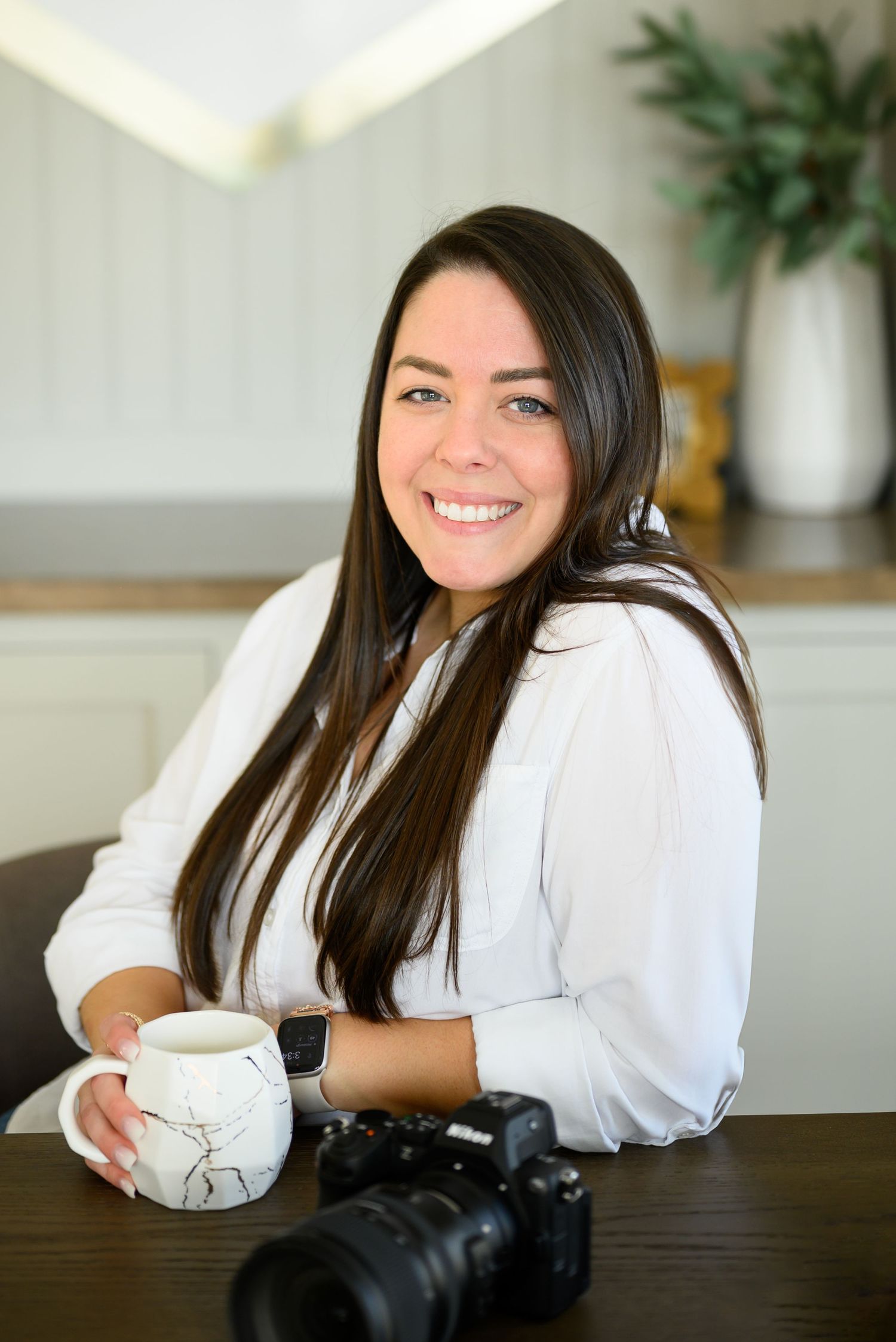 Professional photographer in white shirt sits at dark wooden table with camera and coffee mug in bright modern space.