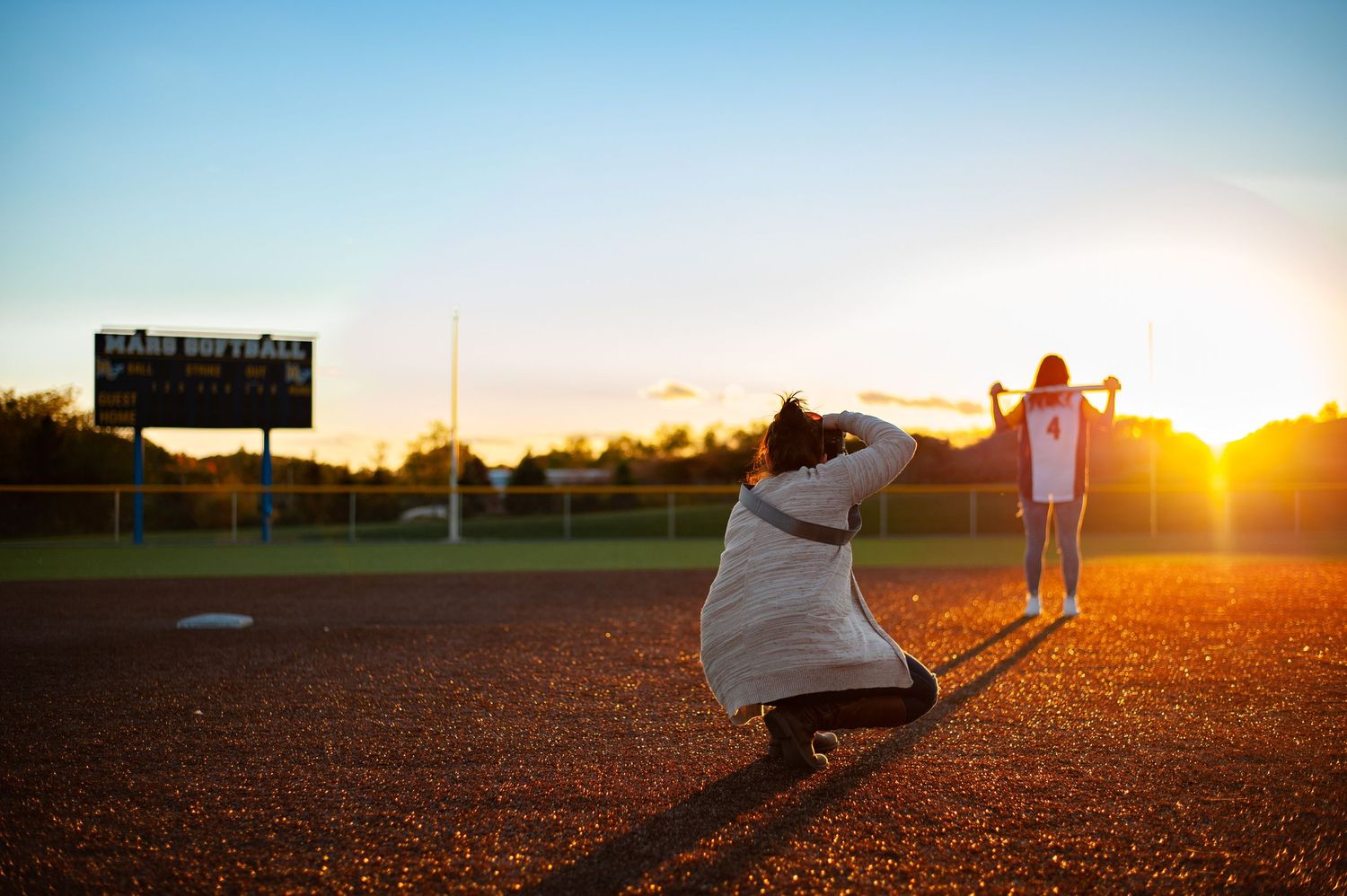 Photographer silhouetted against sunset taking pictures at baseball field.