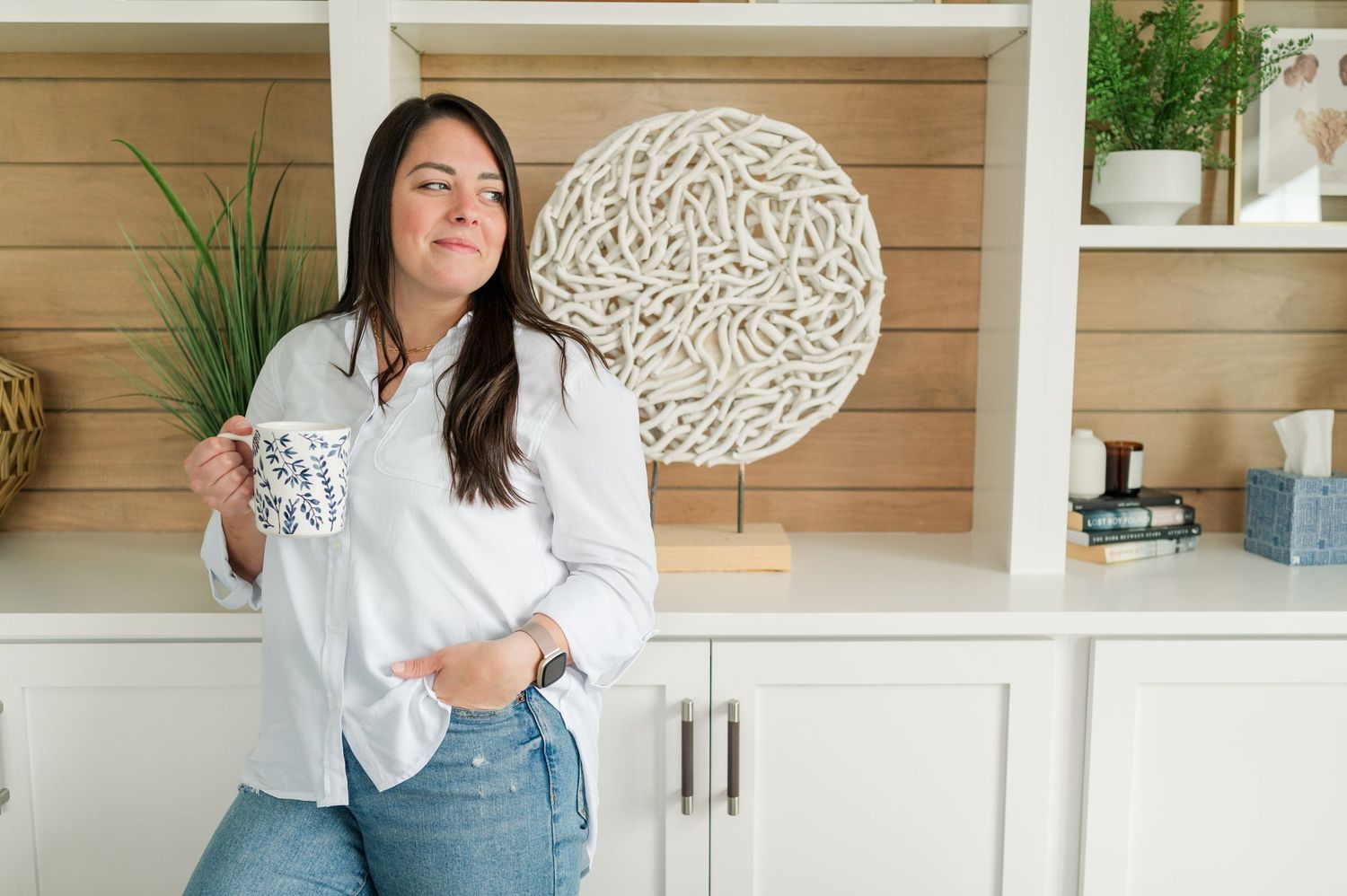Someone in a white button down shirt and jeans stands casually against white cabinets with decorative wall art.