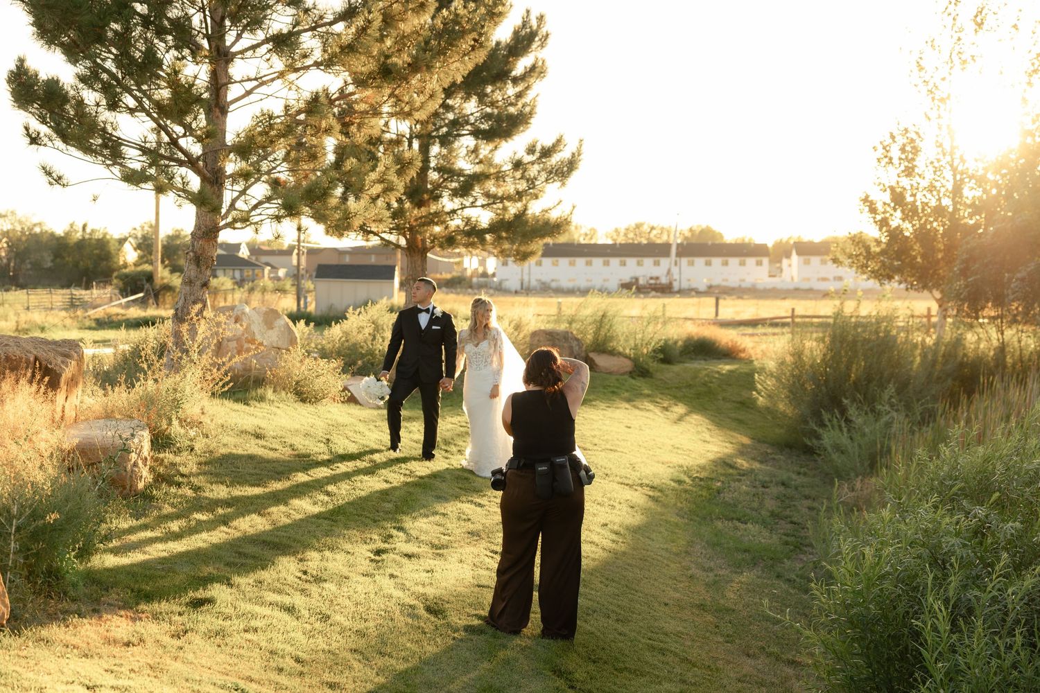 Wedding photographer capturing bride and groom walking on a sunlit grassy path at golden hour.