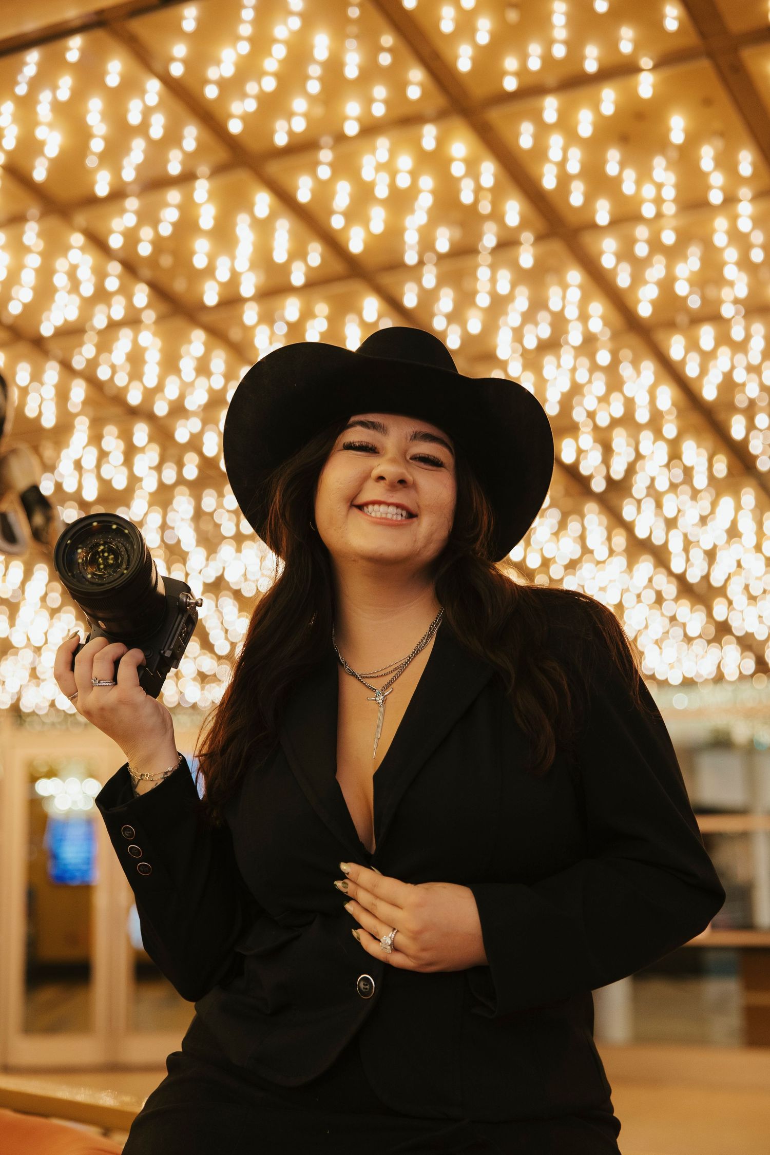 Smiling woman in black blazer and cowboy hat holding a camera with bokeh lights behind her.