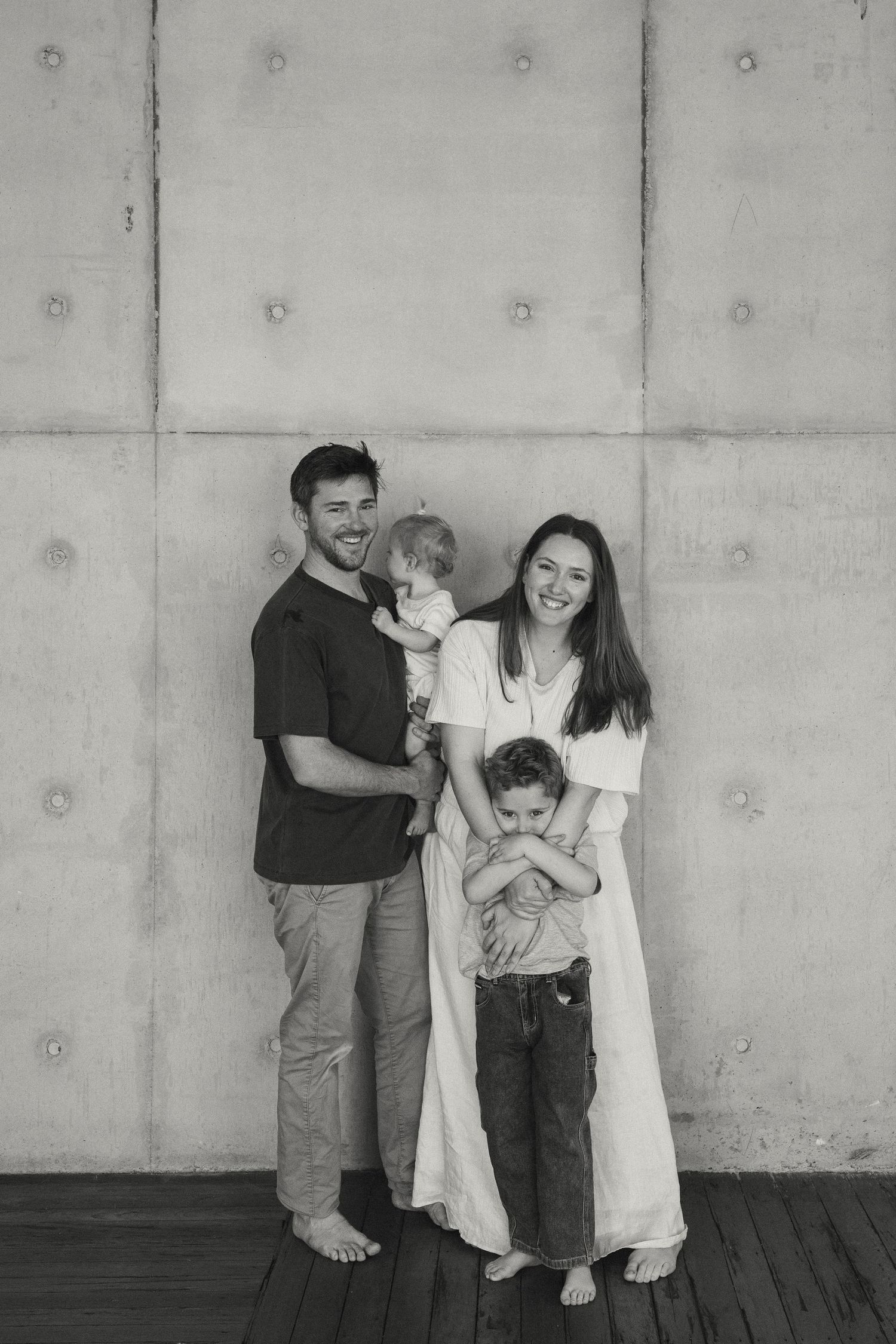 Black and white family portrait of parents with two young children against a concrete wall on a wooden deck.