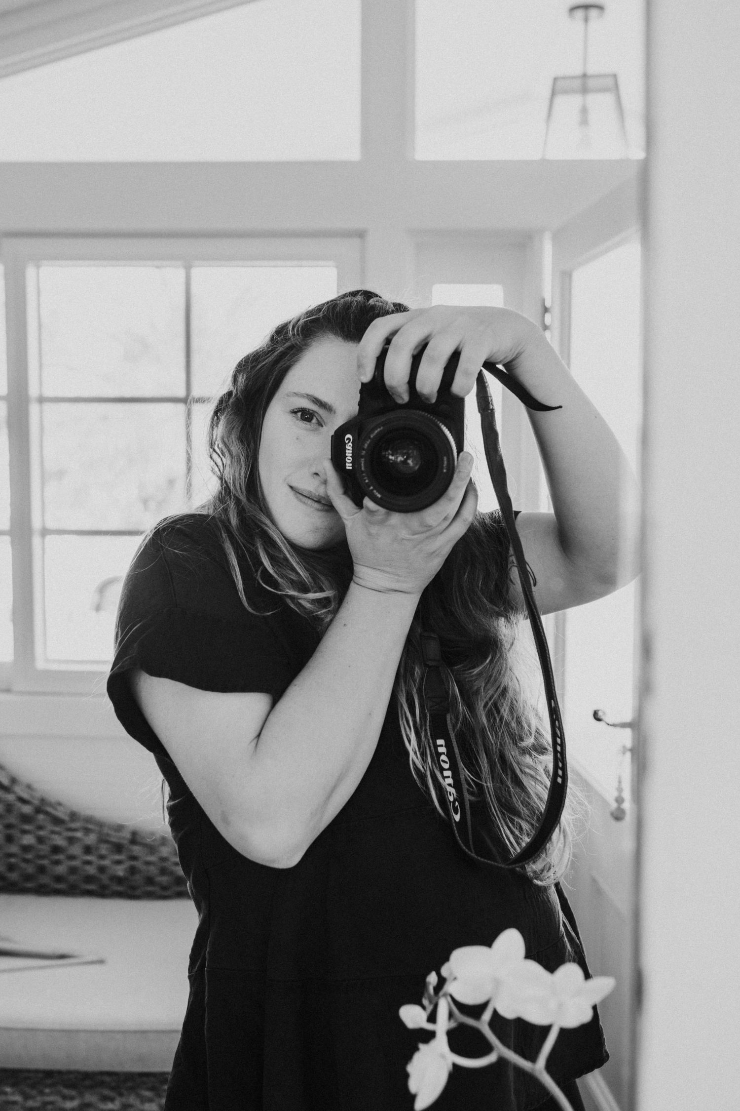 Woman in black t-shirt photographing with Canon DSLR camera indoors, black and white portrait photo.