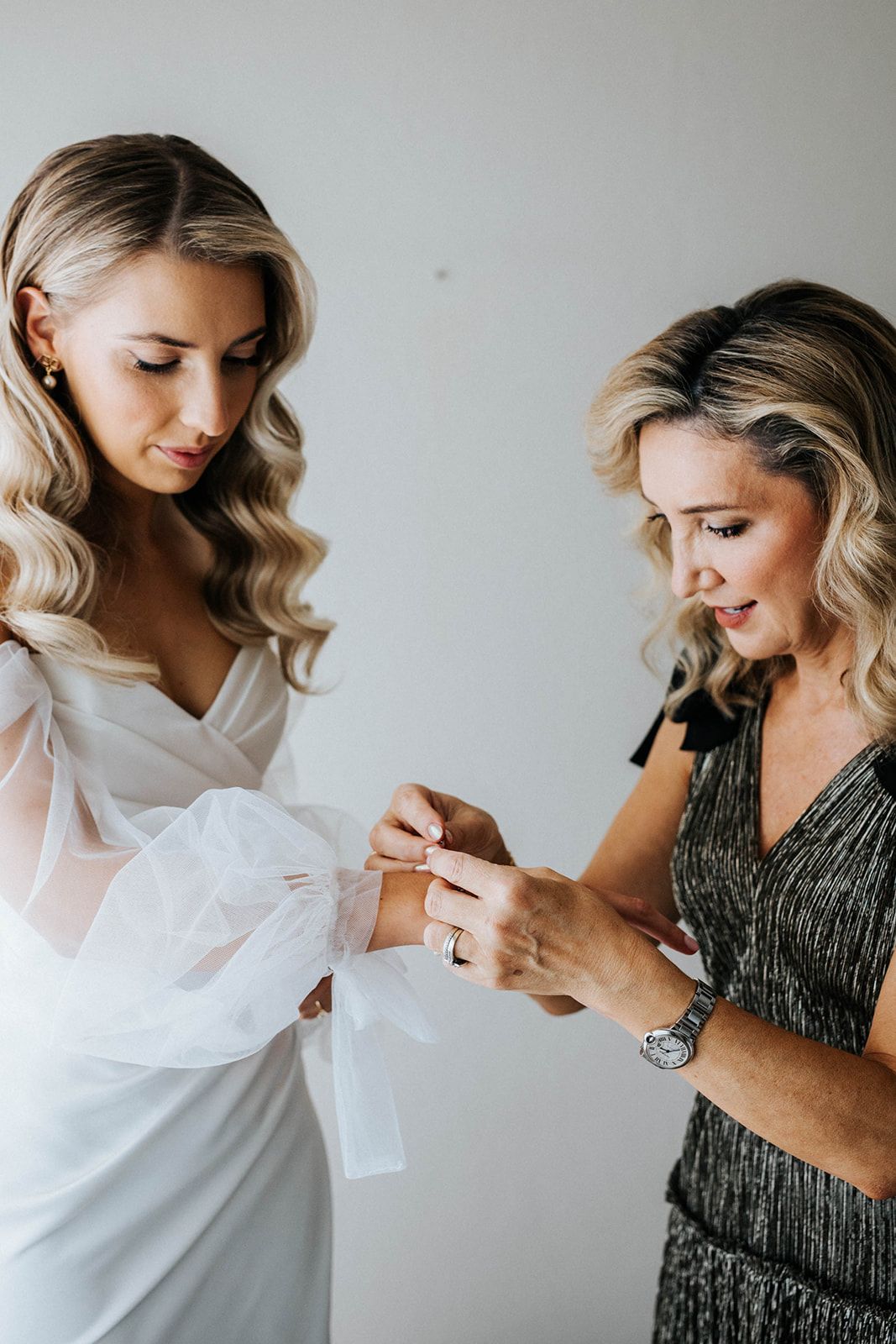 Bride in white gown getting bracelet fastened by woman in silver dress before wedding ceremony.