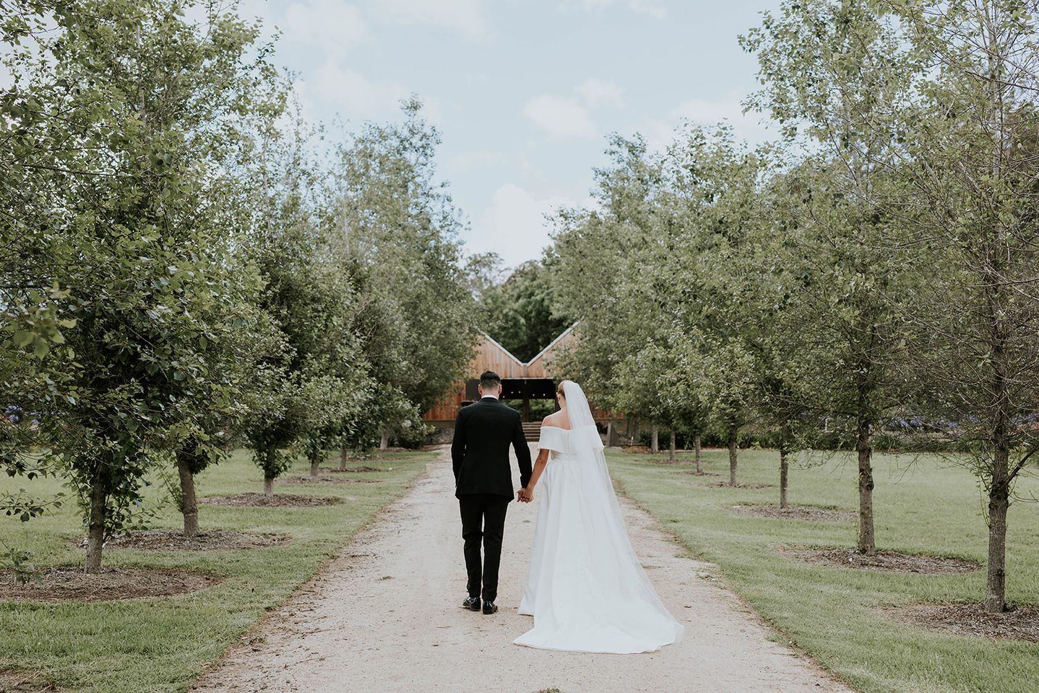 Bride and groom walking hand in hand down a tree-lined path in a lush green garden on their wedding day.