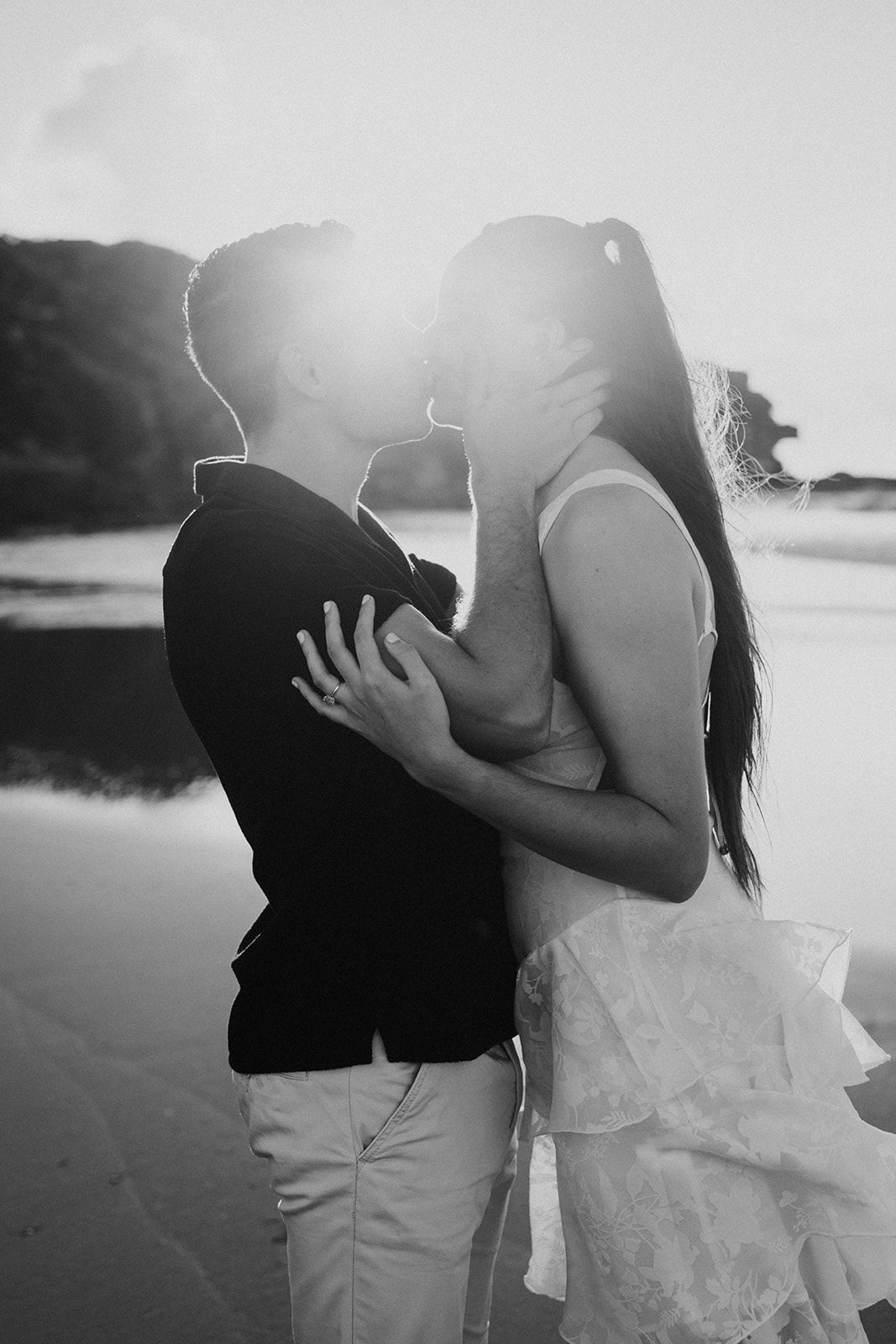 Black and white photo of a couple kissing on a beach at sunset with long hair blowing in the wind.