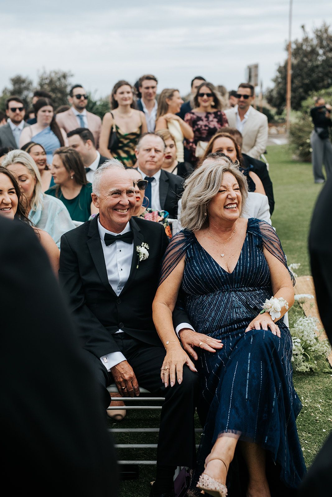 Smiling wedding guests seated outdoors at an elegant ceremony, woman in navy beaded gown with corsage.