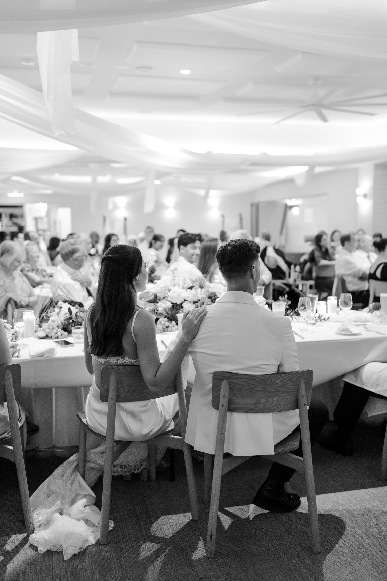 Black and white photo of a couple seated at a wedding reception table, surrounded by guests and floral centerpieces.