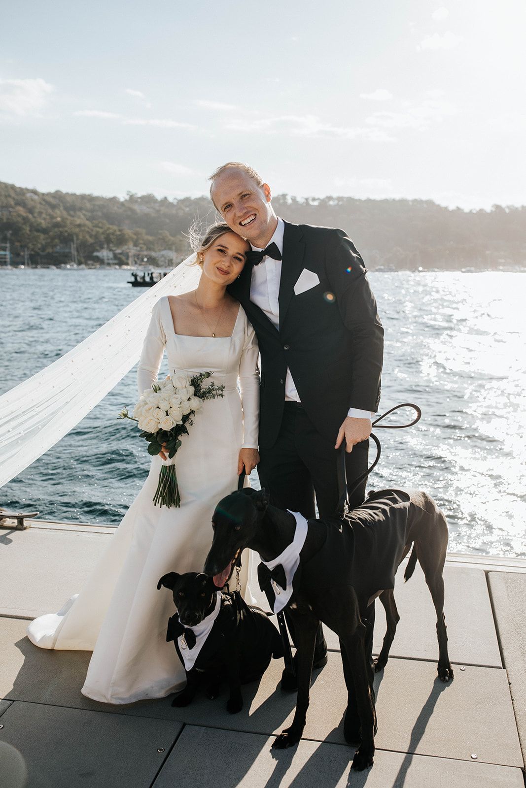 Bride and groom posing with two black dogs on a waterfront dock, bride holding white bouquet in elegant long-sleeve gown.