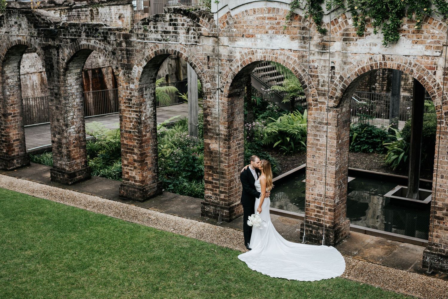 Bride and groom share romantic moment among historic brick arches and lush greenery at elegant outdoor wedding venue.