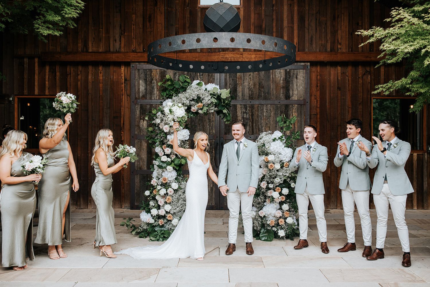 Bride and groom celebrate with wedding party outside rustic barn venue, surrounded by lush floral arch arrangements.