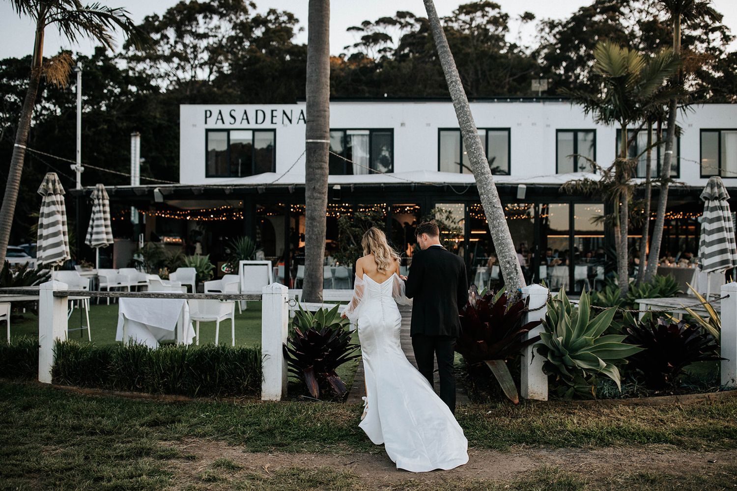 Bride and groom stand before Pasadena venue with outdoor dining, palm trees, and string lights at their wedding.