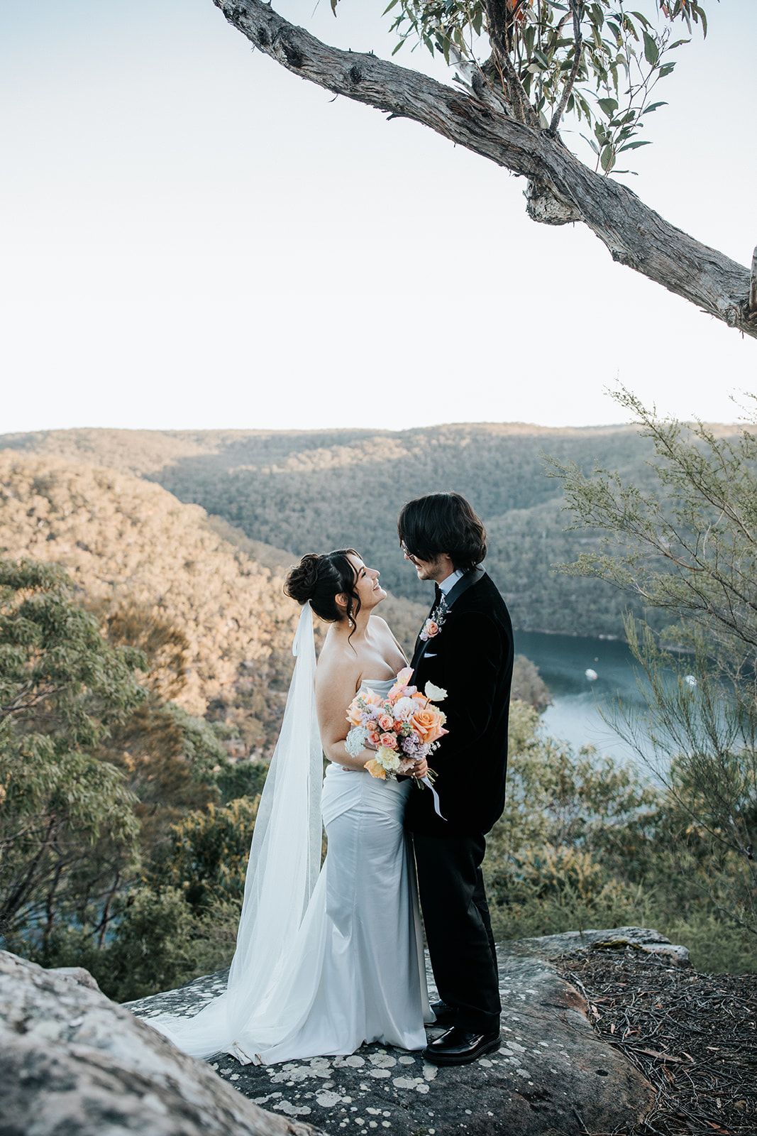 Bride and groom share a romantic moment on a rocky cliff overlooking a scenic river valley and forested hills.