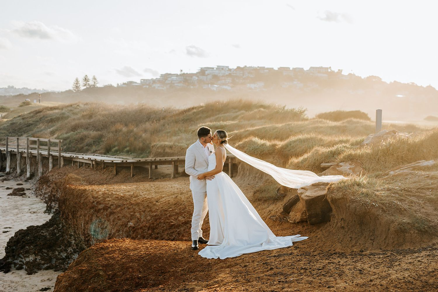 Bride and groom embrace on a coastal dune at golden hour, veil flowing in the wind with misty hills behind.