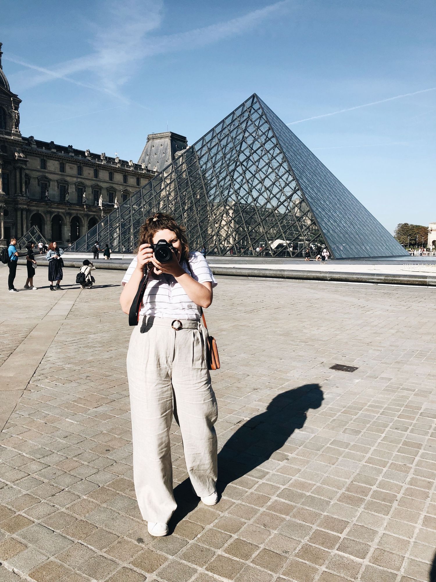 Woman photographing at the Louvre Museum pyramid in Paris, wearing linen wide-leg pants on a sunny day.