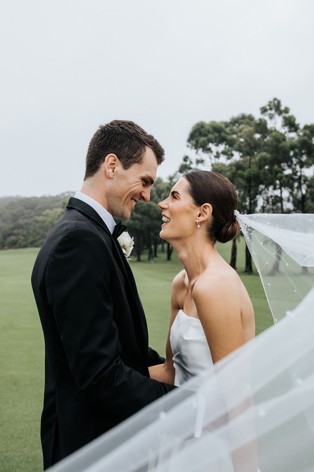 A bride and groom share a romantic moment outdoors on a golf course, smiling at each other while her veil flows in the breeze.